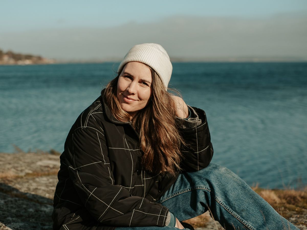 Kvinna som sitter på ett berg vi havet, hon tittar in i kameran.