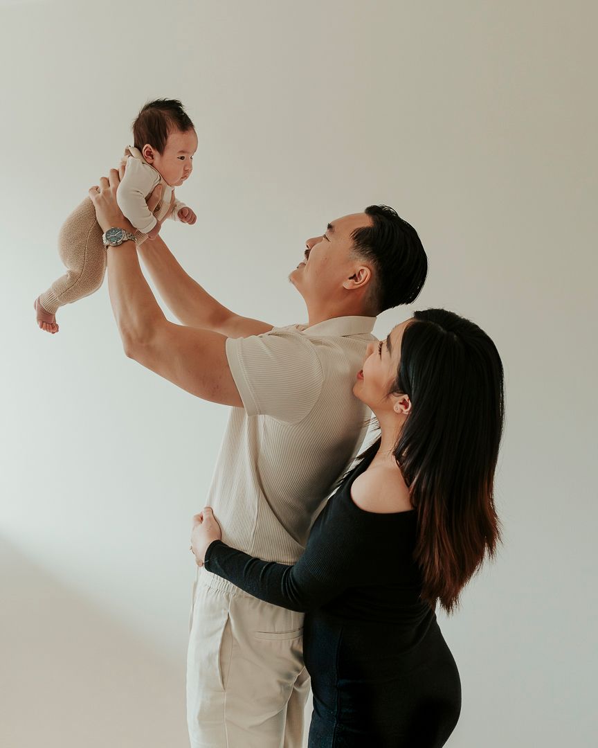 Familjefotografering i hemmet i Göteborg. Pappan håller upp bebisen i luften och mammans står och kramar honom bakifrån