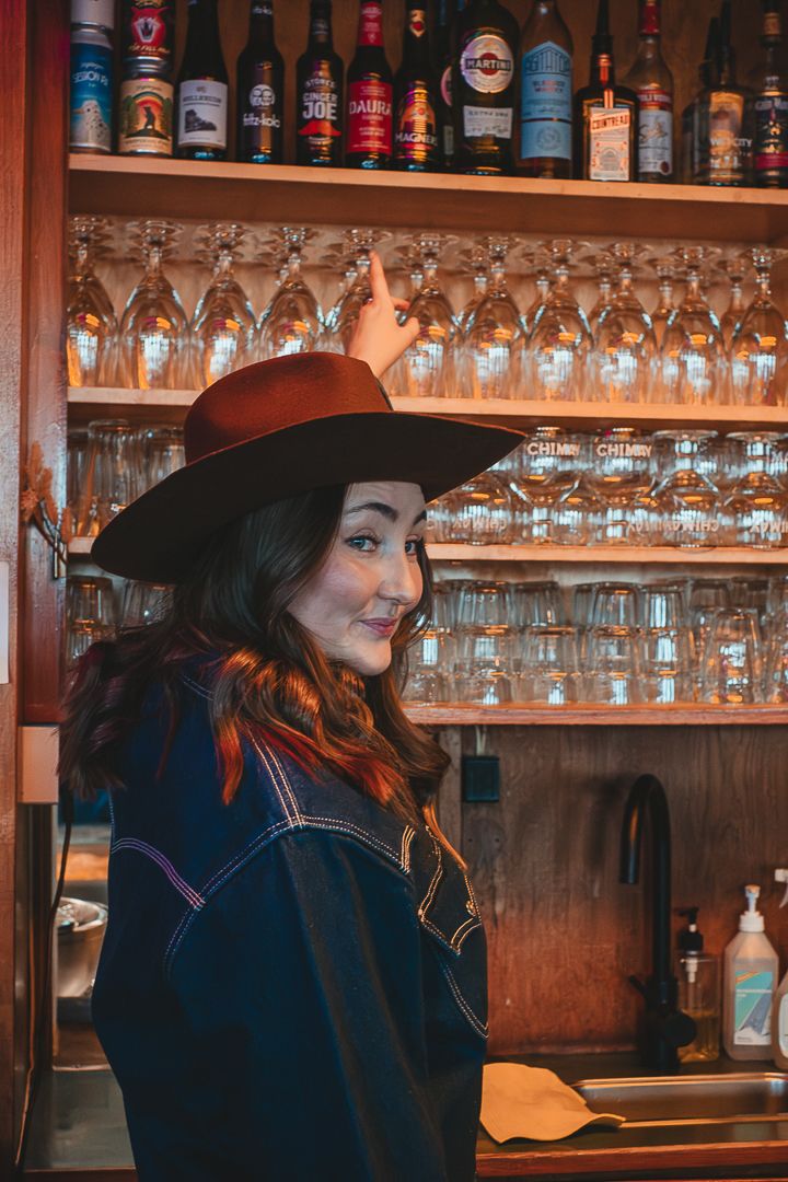 En kvinna i brun cowboy hat som står i en bar, hon sträcker upp handen för att ta ner ett glas.