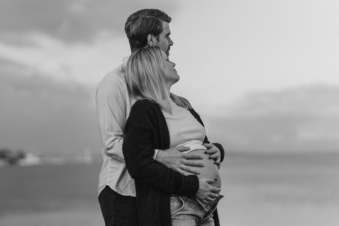 Par som som står och håller om varandra i havet, de blickar ut. Kvinnan väntar barn och står lutad mot mannen. De har händerna på magen
