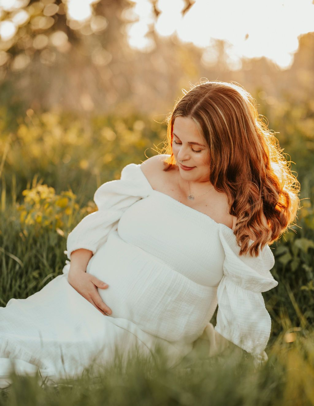 En gravid kvinna som sitter på en äng i solskenet i en vit klänning. Sitter på en äng i Kungsbacka med handen på magen.