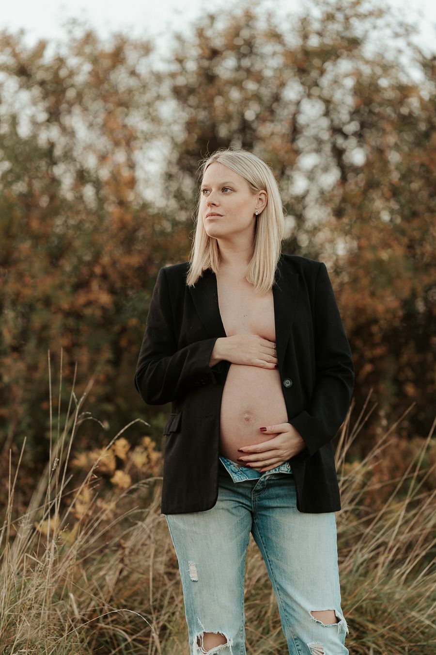 Gravid kvinna som står med en öppen kavajjacka och jeans på en äng i solnedgången i Kungsbacka. Hon har händerna på magen.