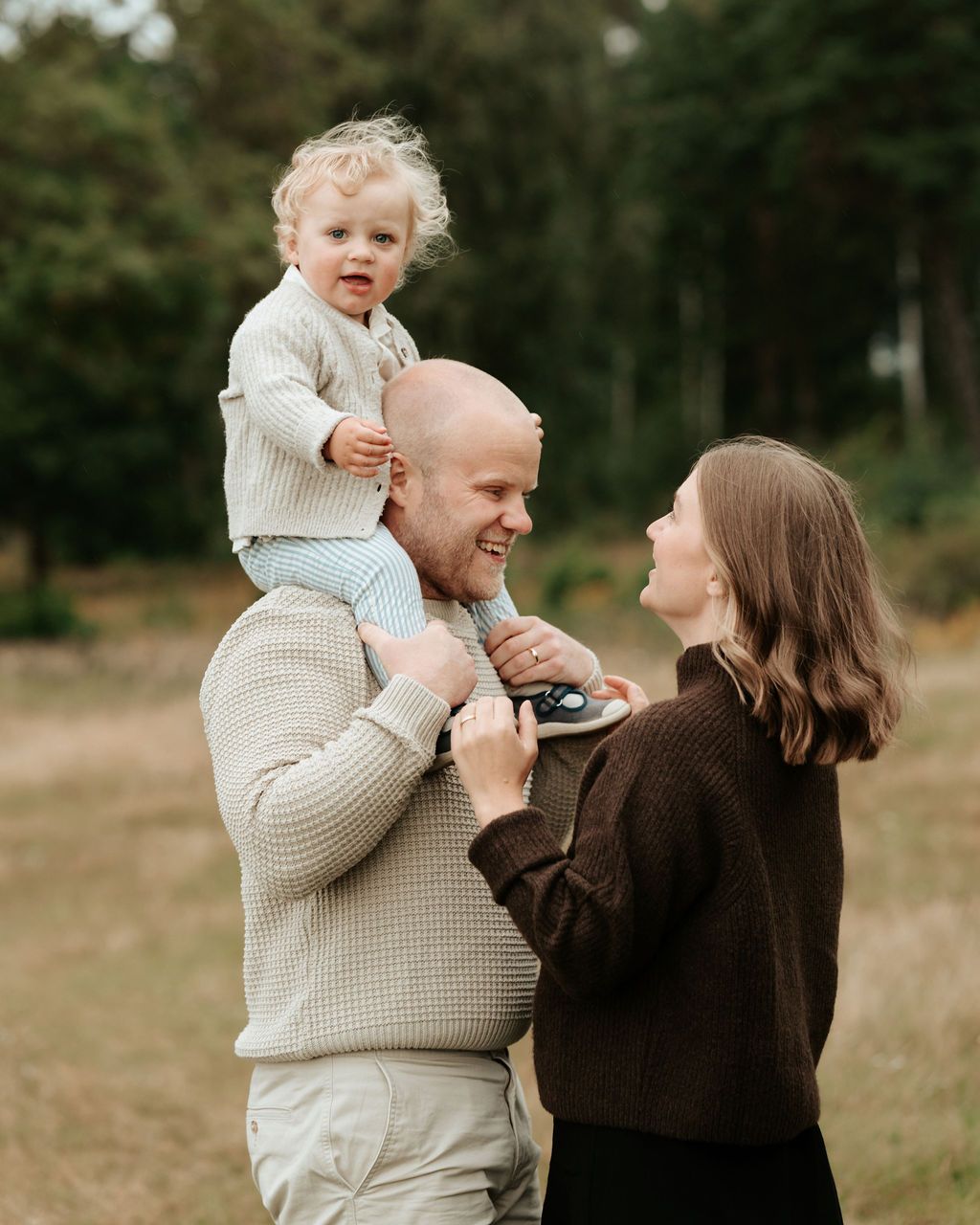 Familjefotografering på en äng i Kungsbacka, barnet sitter på pappans axlar och mamman står framför dom och håller i barnens fötter