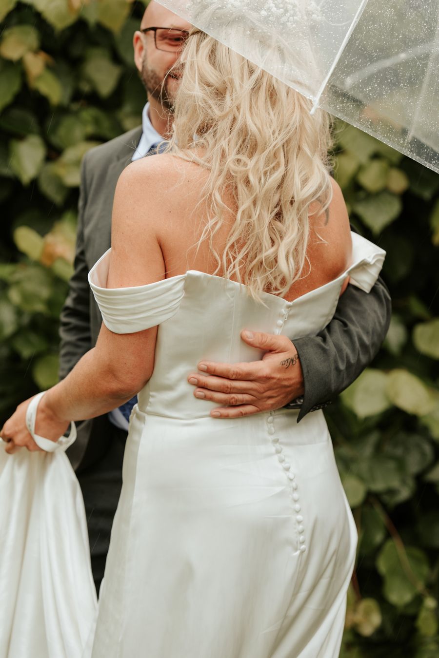 Brudpar som står i regnet under ett paraply. Man ser ryggen på brudens klänning och mannen håller om henne.