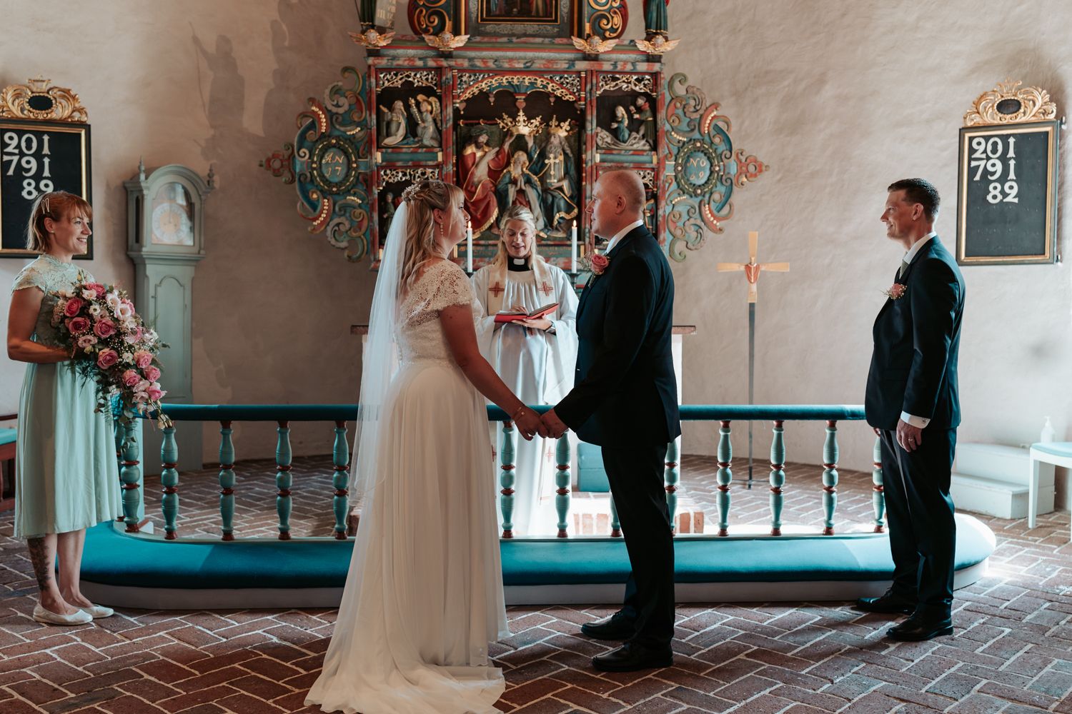 Brudparet står i kyrkan under vigseln, de håller varandra i händerna medans prästen pratar.