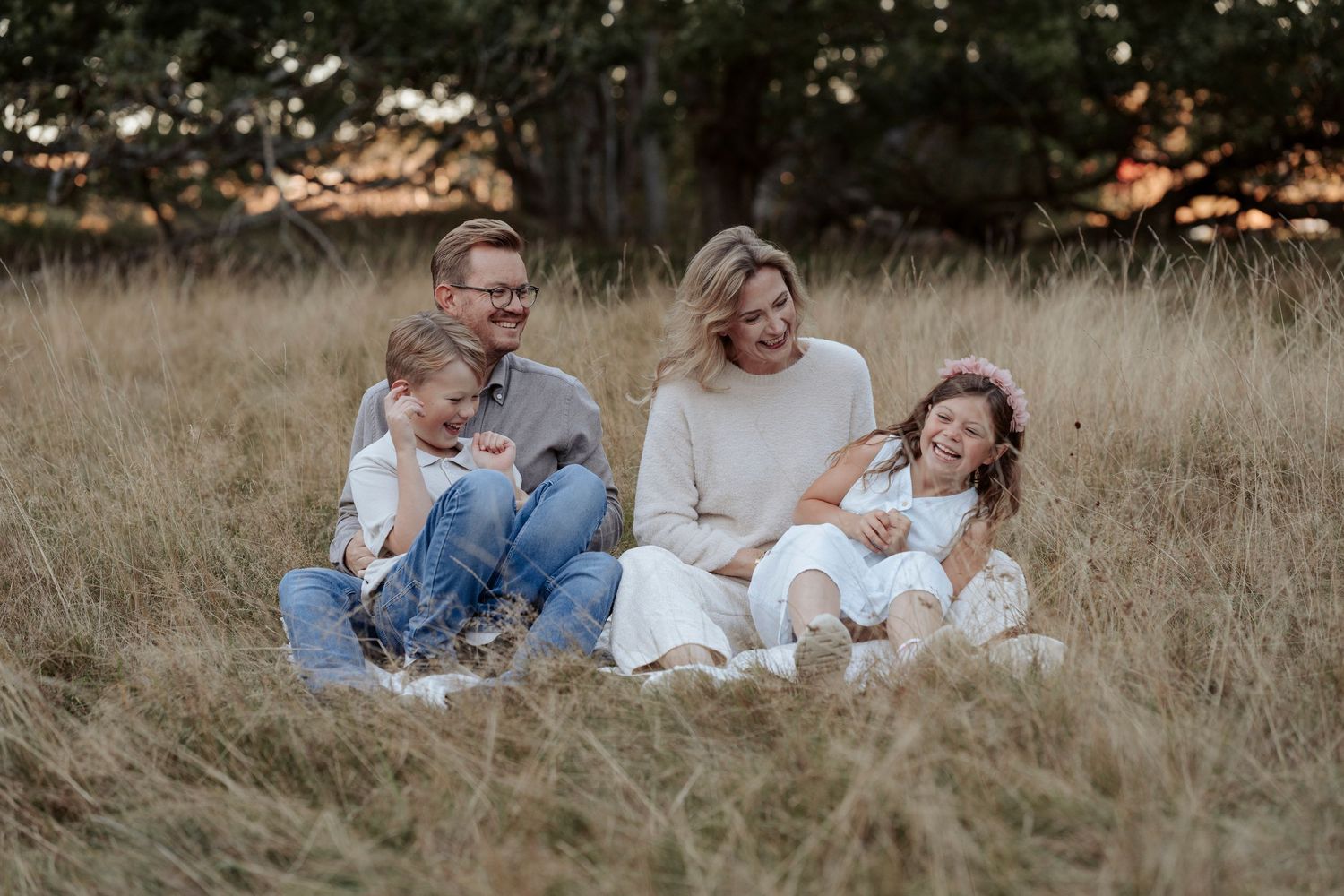 Familj som sitter och håller om varandra på en filt på en äng i solnedgången. Föräldrarna kittlar barnen som skrattar.