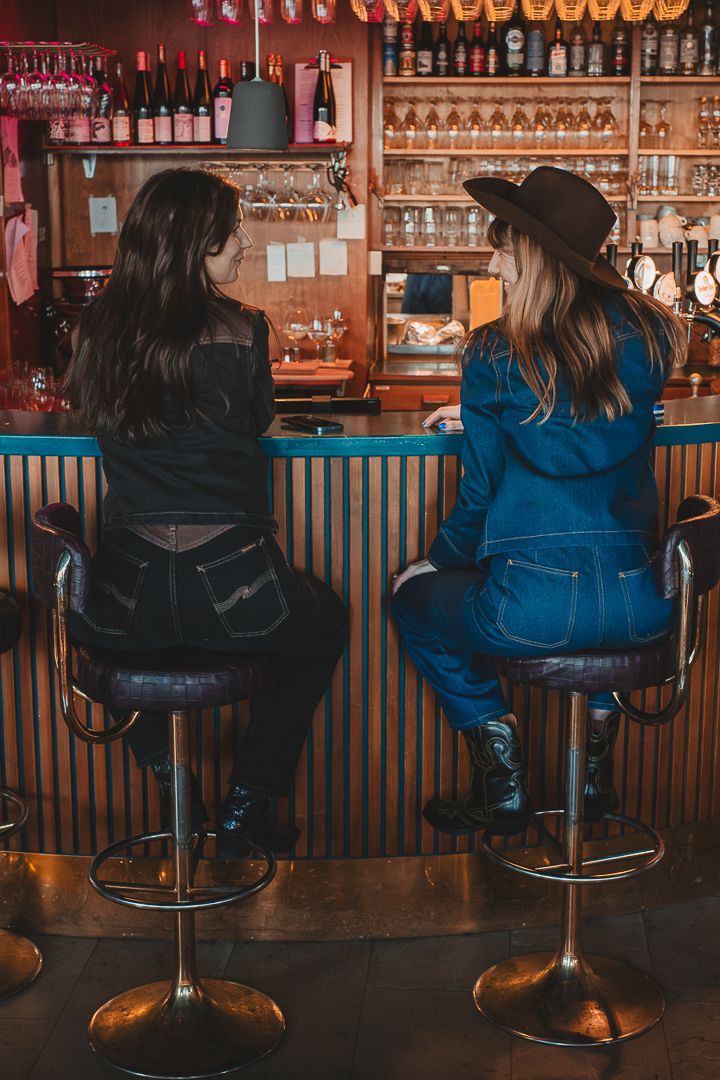 Två kvinnor i jeanskläder sitter i en bar på varsin barstol, de har ryggarna mot kameran. Den ena har på sig en cowboy hat.