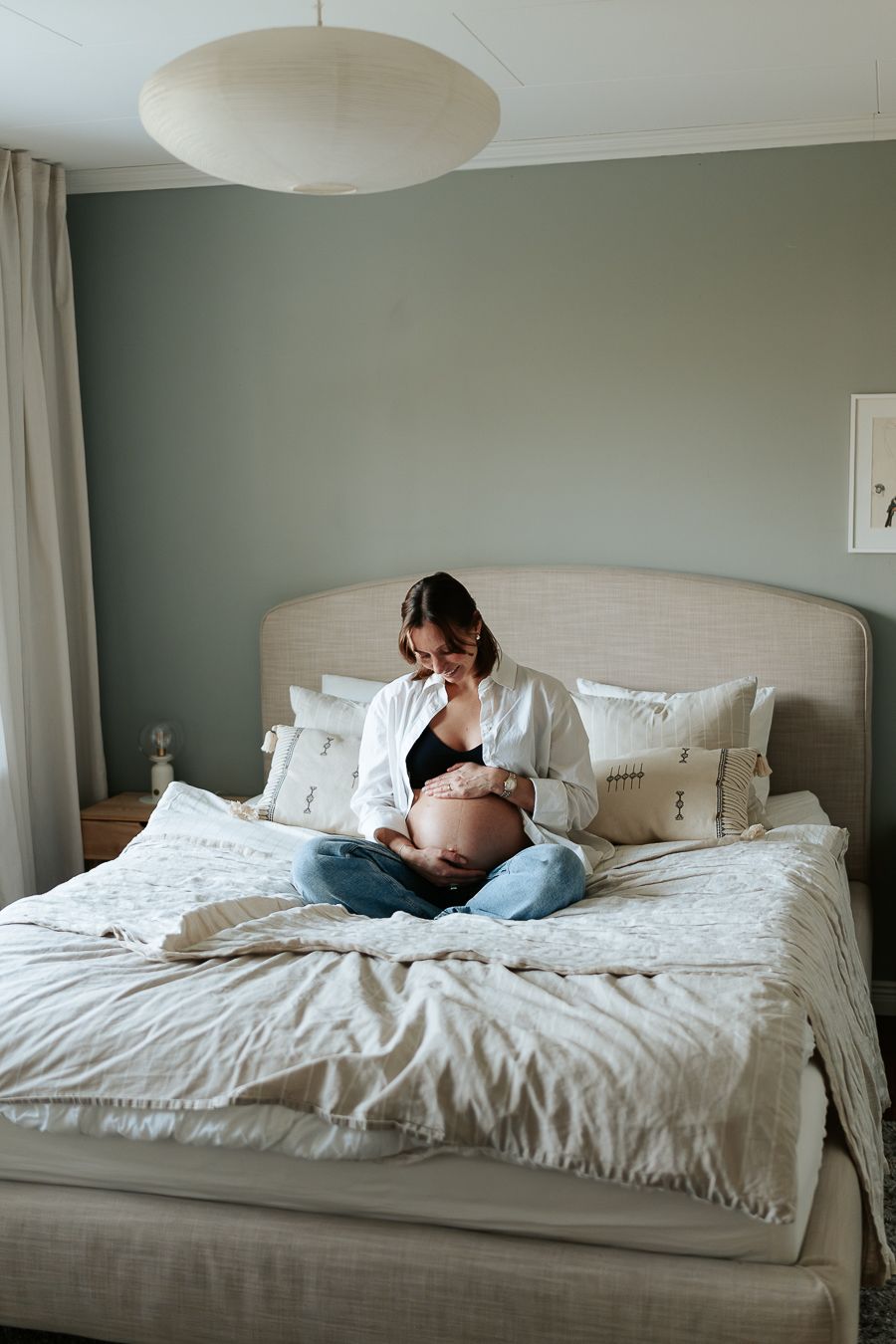 En kvinna sitter på en säng i ett sovrum. Hon är gravid och har händerna på sin bara mage och tittar ner på den