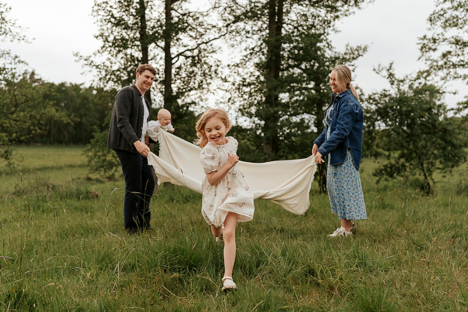 Två föräldrar håller i en filt som ett barn springer under. En härlig busig stämning i en grön miljö.