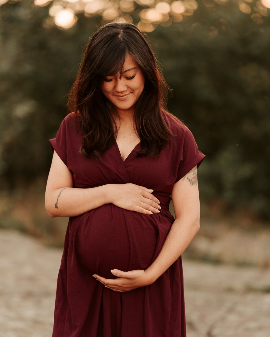 Gravid kvinna står i en lila klänning i solnedgången och håller båda sina händer om sin mage.