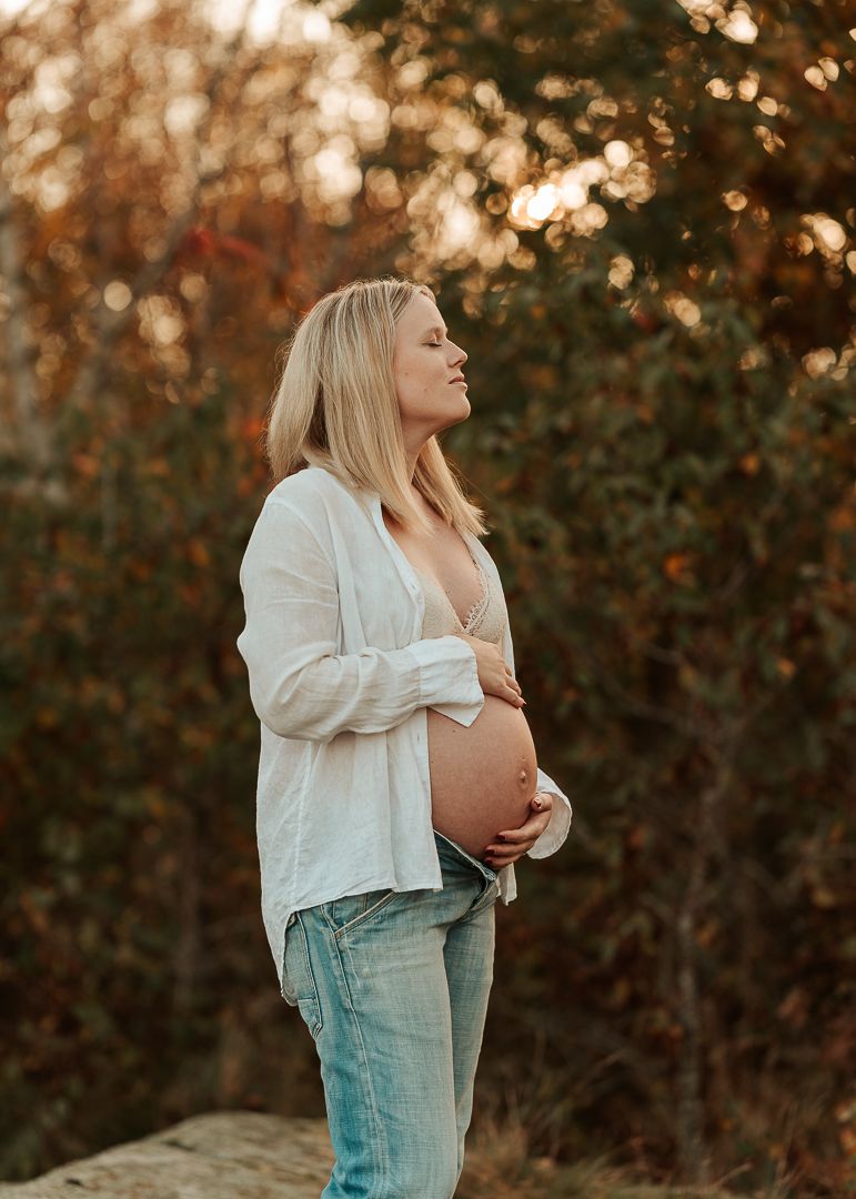 Kvinna som är gravid står framför grönska där man kan se solens strålar bakom. Hon har på sig en uppknäppt skjorta och har händerna på magen