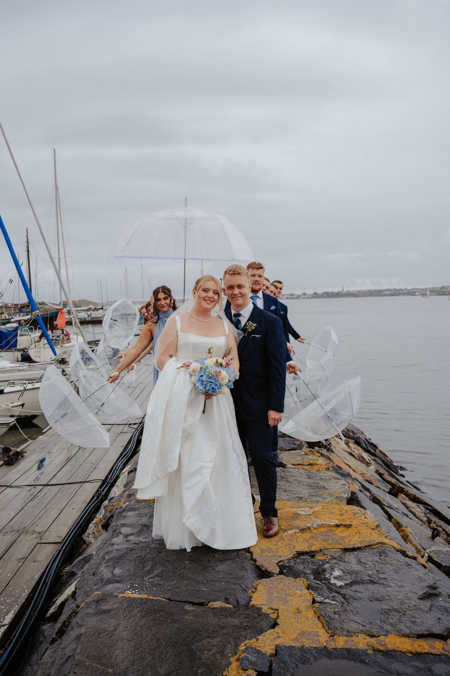 Brudpar och brudfölje som står på en kaj i regnet. Brudföljet står bakom brudparet och lutar sig fram. De ha paraplyer.