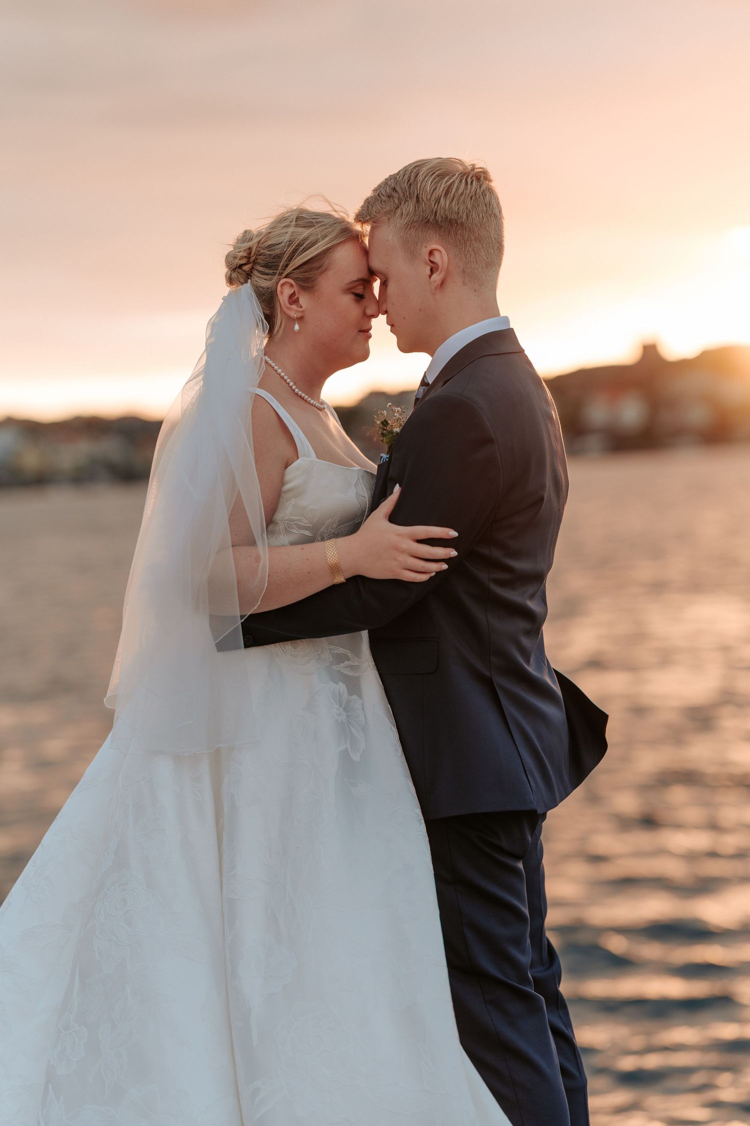 Ett bröllopspar som står panna mot panna vid havet i solnedgången.