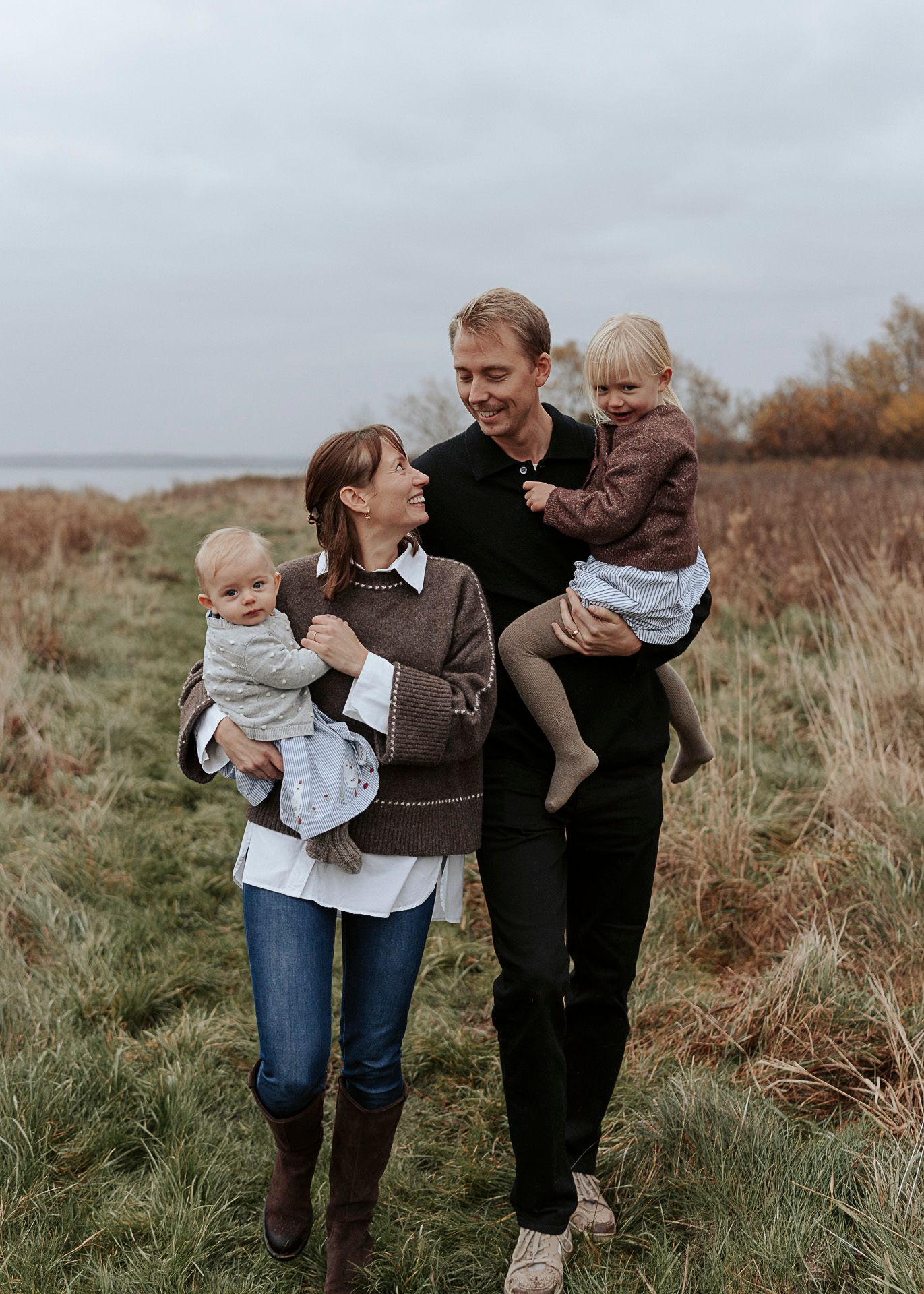 Två föräldrar håller varsitt barn i famnen medans de går på en äng. De tittar på varandra. Höstig miljö.