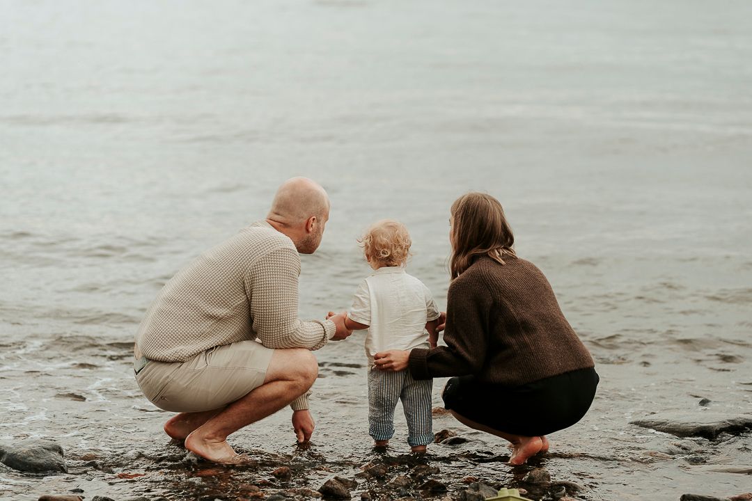 En pappa, mamma och son sitter på huk vid strandkanten i Kungsbacka. De har ryggarna mot kameran och kollar ut över havet.