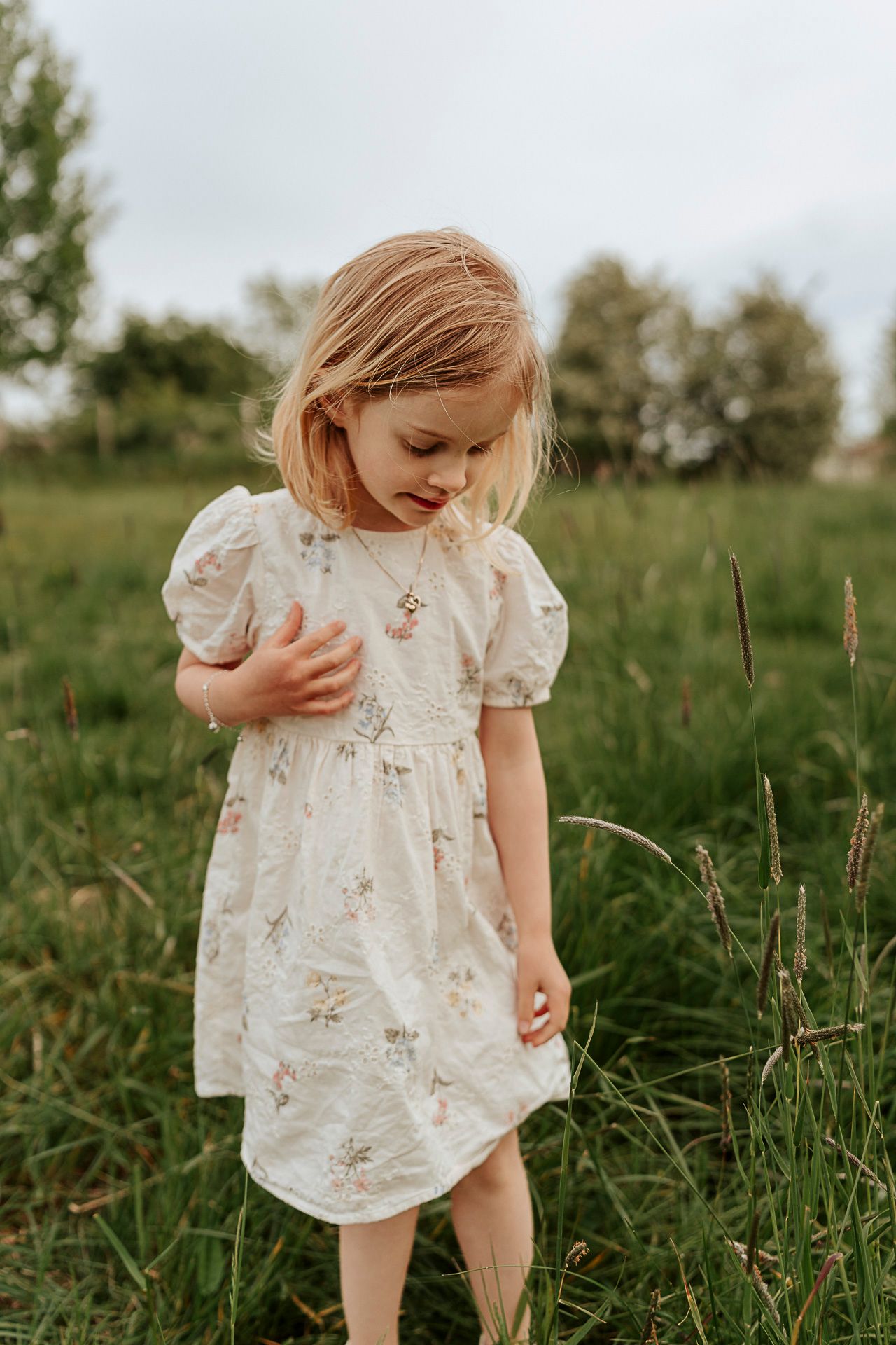 En flicka i en vit klänning med blommor på som står utomhus. Hon tittar ner mot marken.