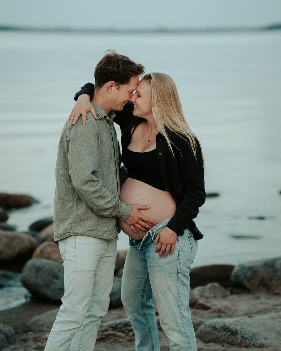 Par som väntar barn står vid en strand vid havet. De har ansiktena mot varandra och lutar sina pannor ihop. Kvinnan är havande
