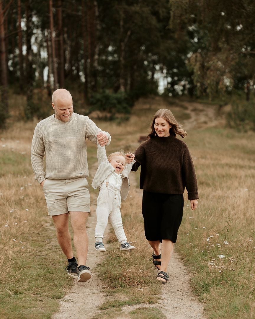En familjefotografering vid en äng i Kungsbacka. En mamma och pappa håller sitt barn i handen och svingar honom i luften.