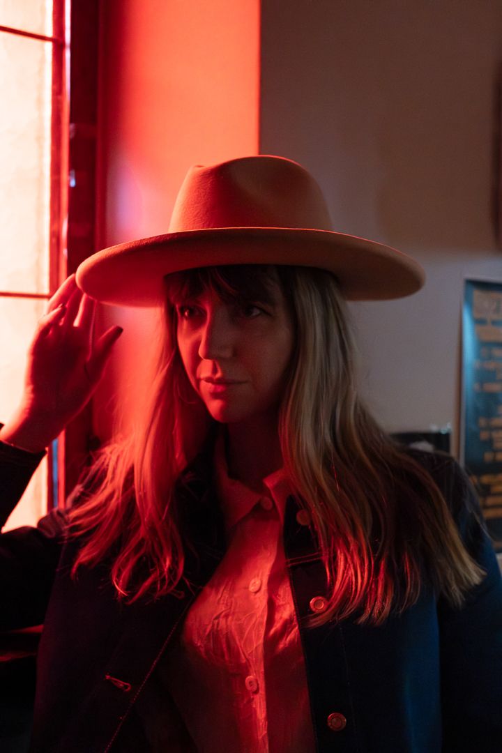 ett porträttfoto taget i rött ljus på en kvinna som har på sig cowboy hat. Hon står vid ett fönster och håller ena handen på hatten.