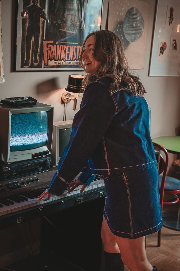 En kvinna som har på sig jeansjacka och jeanskjol står vid ett piano