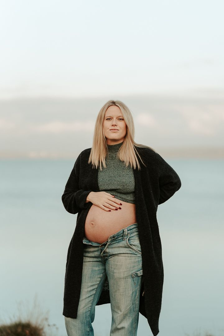 Gravid kvinna står vid havet och tittar rakt in i kameran medans hon håller en hand på sin gravida mage. Magen är bar