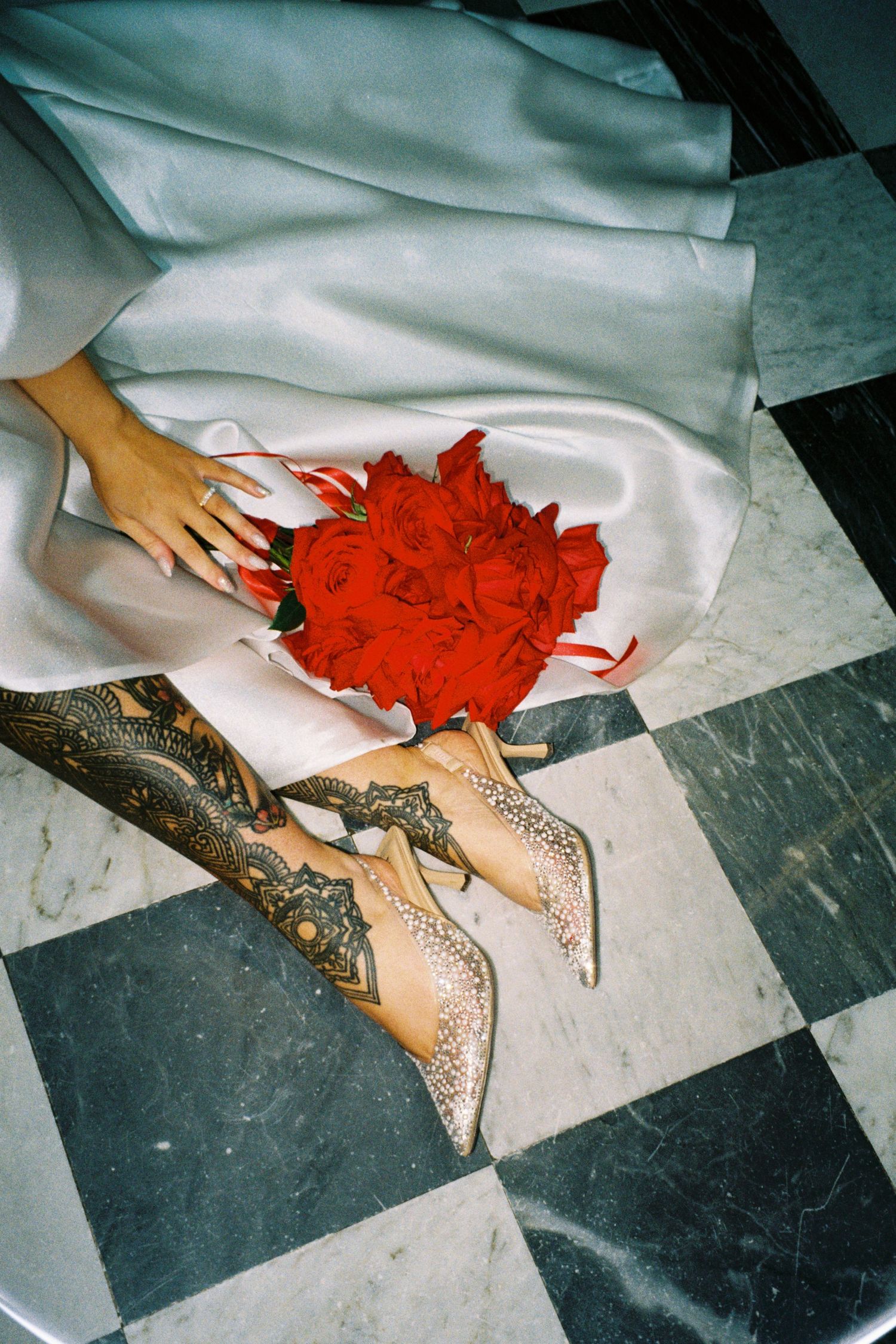 A person wearing a white dress and henna-decorated feet holds a vibrant red bouquet on a checkered floor.