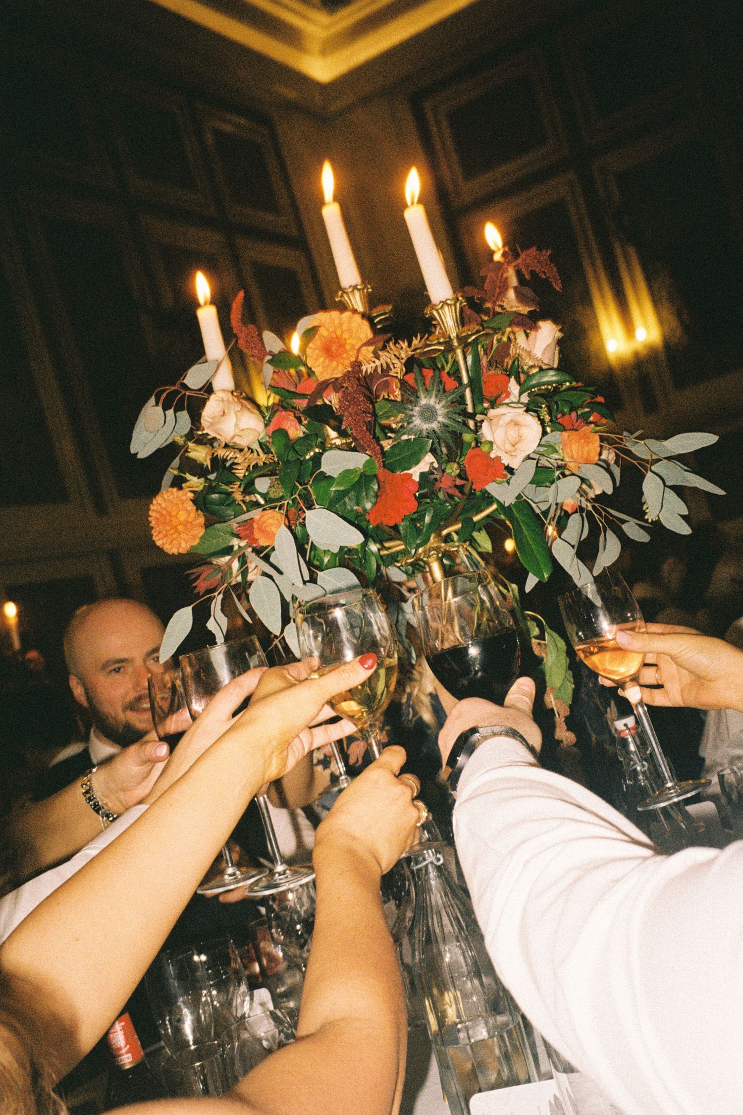 Multiple wine glasses clink together in celebration, adorned with floral arrangements and candlelight.