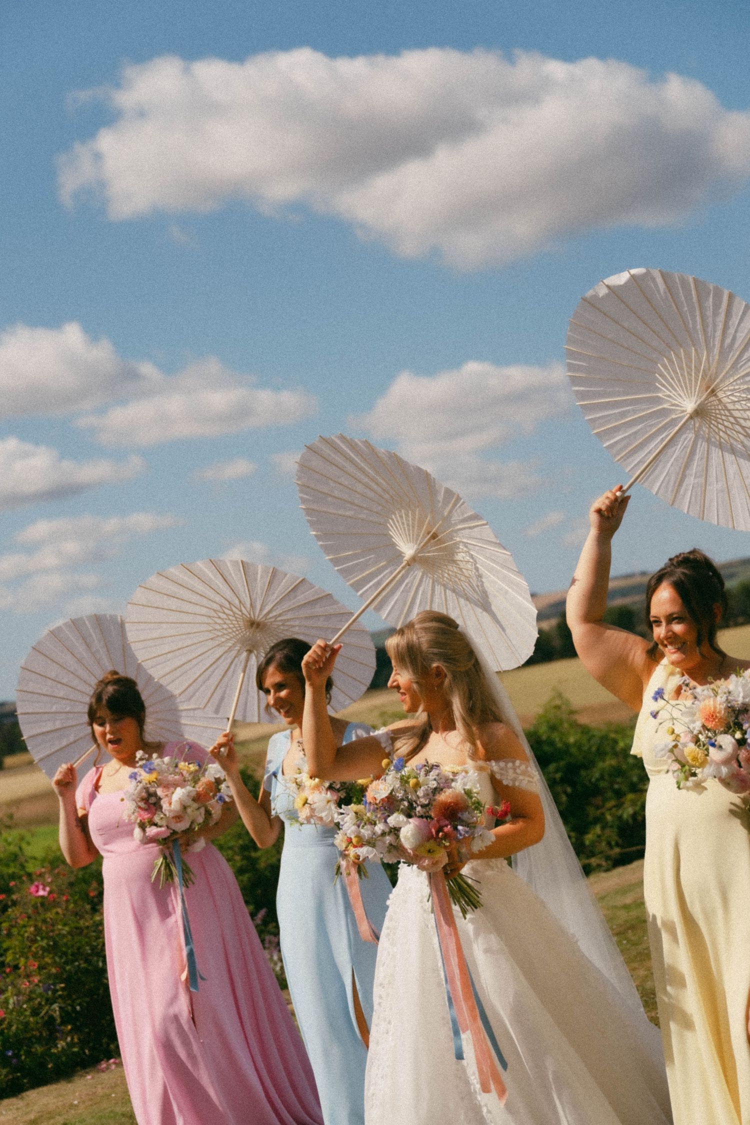 A group holding white paper parasols in coordinated pastel bridesmaid dresses pose together against a blue sky backdrop.
