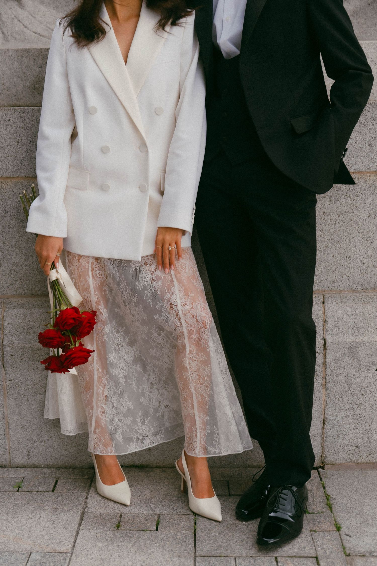 A couple sits on stone steps with white formal wear and a bouquet of red roses creating a romantic moment.