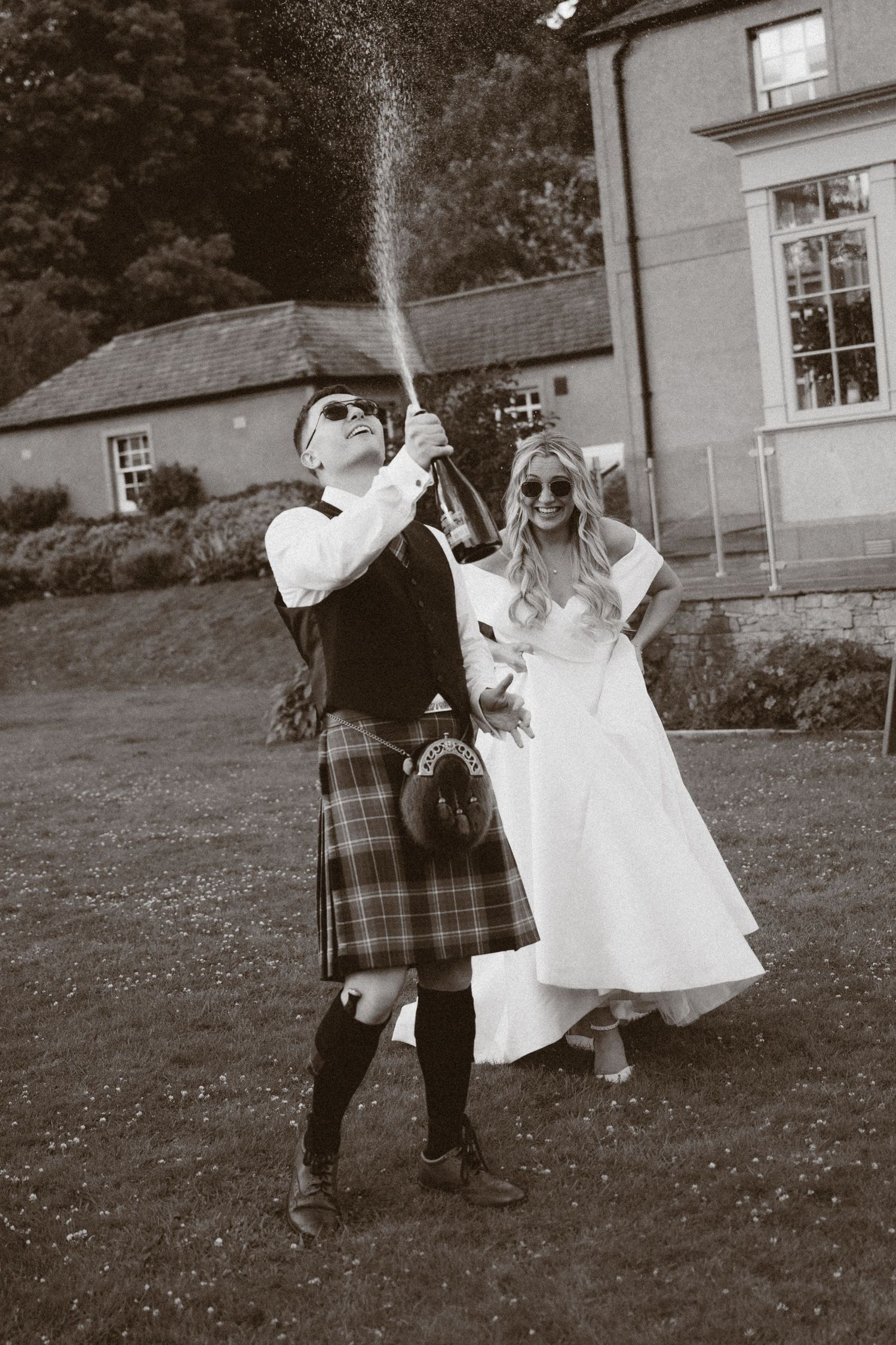 Groom in tartan kilt pops champagne bottle outdoors while bride in white dress laughs beside him at Scottish wedding.