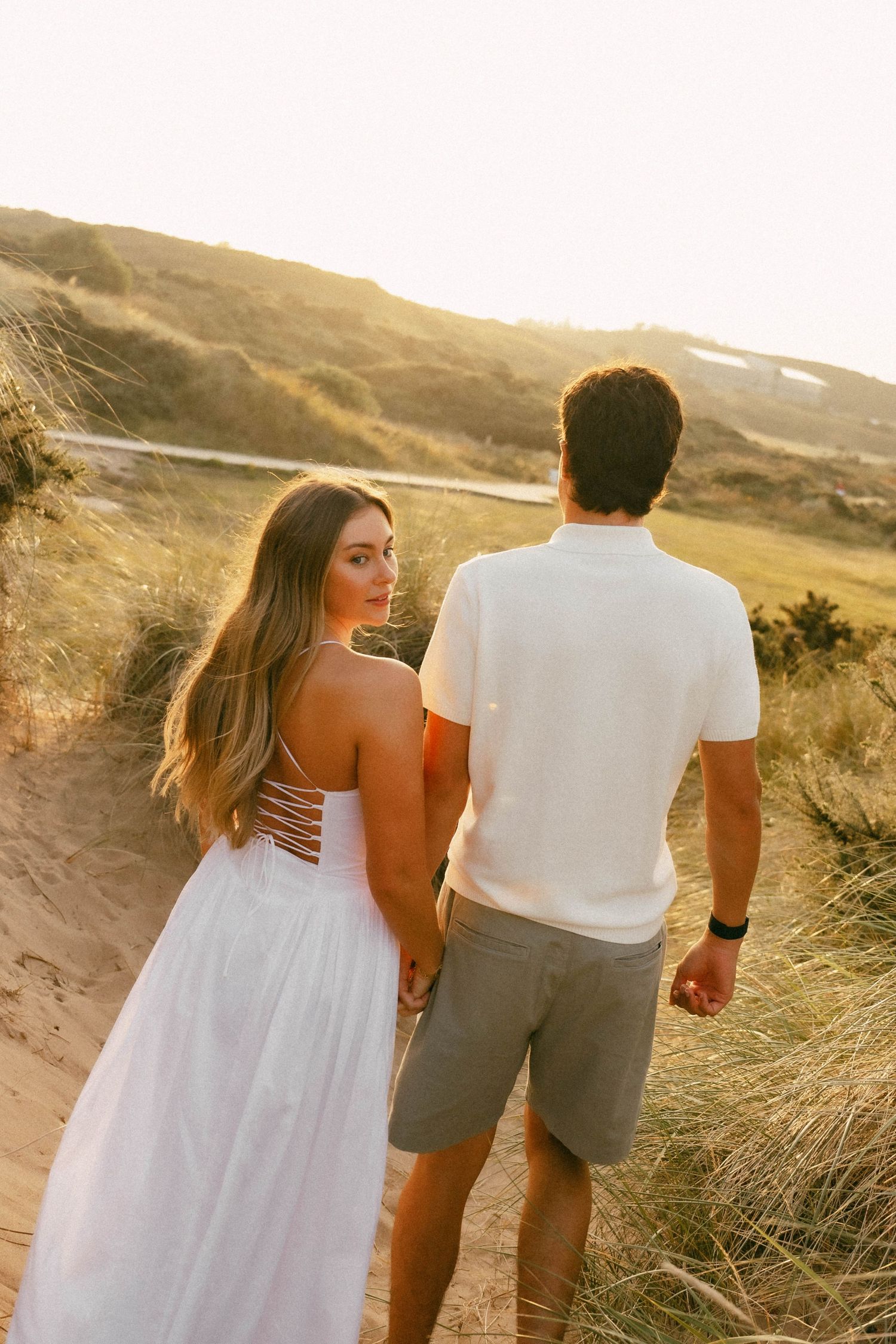 Couple holding hands on sandy beach dunes at golden hour, woman in white dress looking back over shoulder.