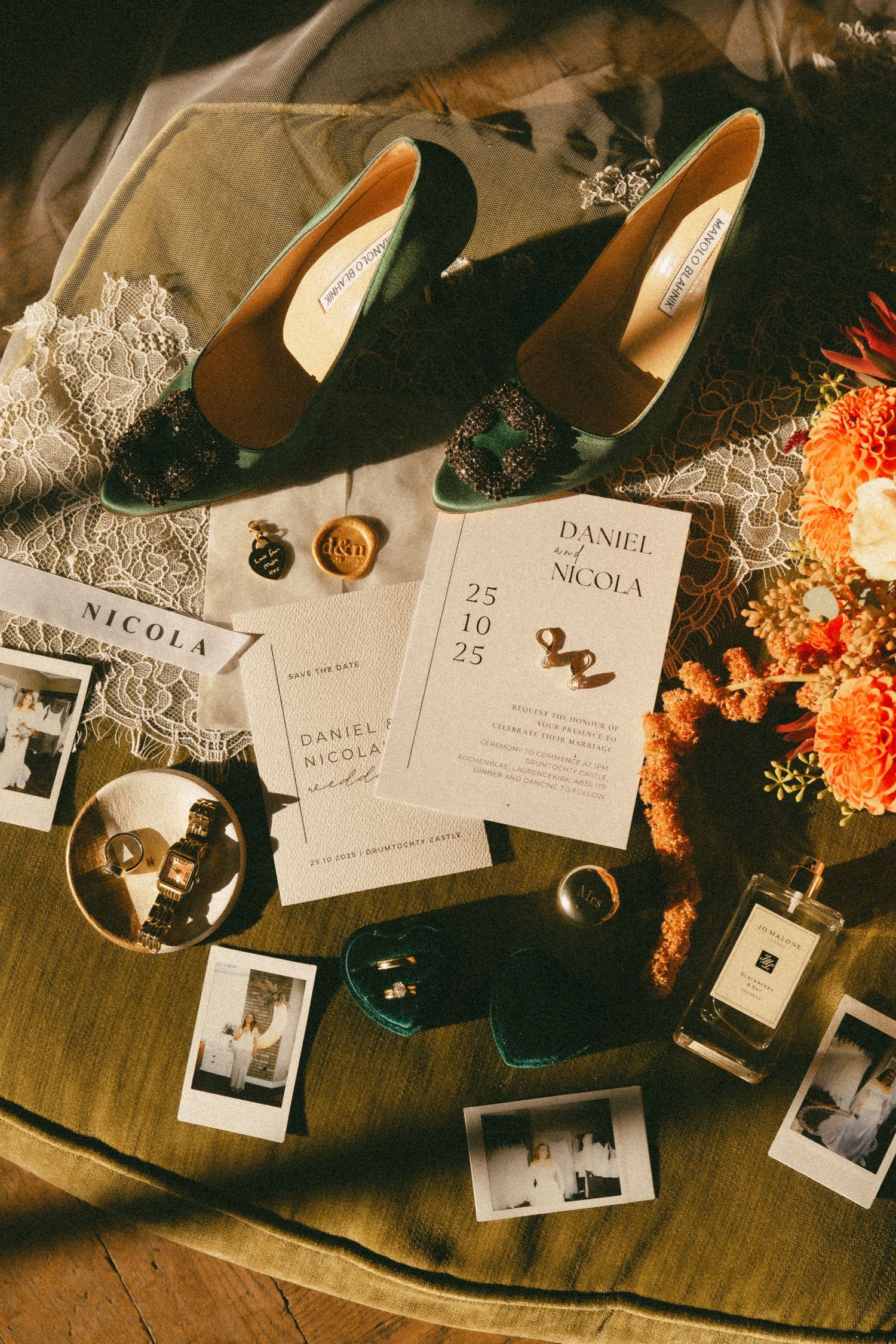 Wedding details flatlay with black heels, invitation cards, wax seals, polaroid photos and orange flowers on wooden surface.