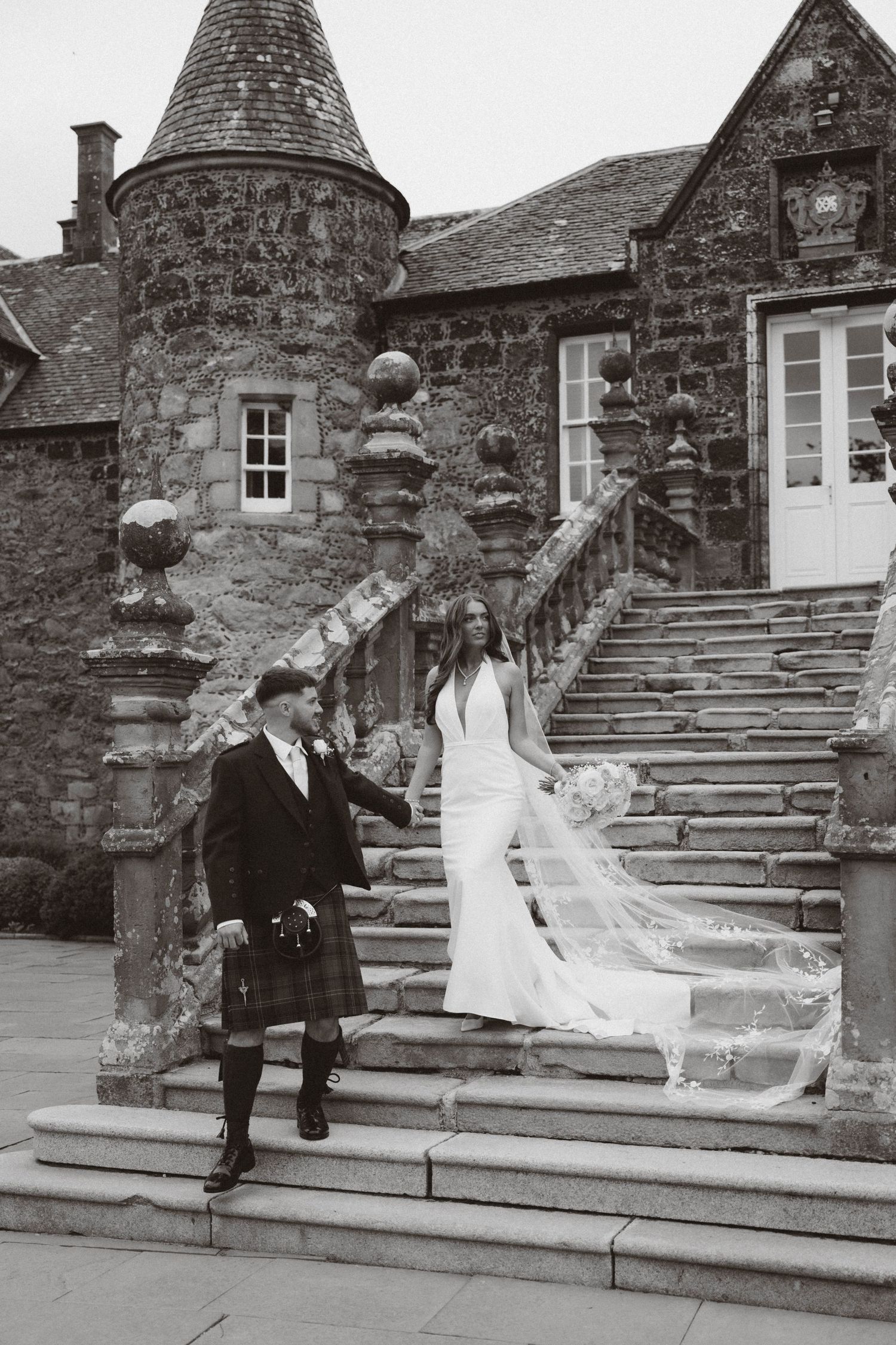 A couple in traditional Scottish wedding attire pose on stone steps outside a historic stone castle with a turret.