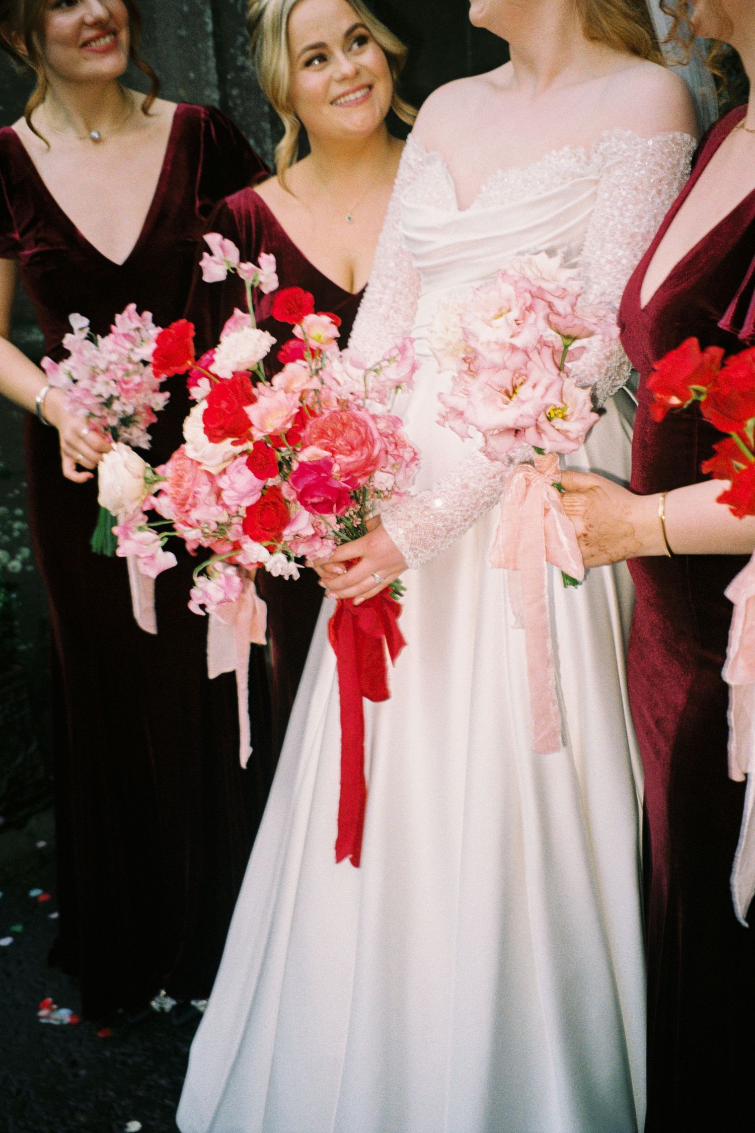 Wedding party members in burgundy and white dresses hold pink and red floral bouquets tied with ribbons.