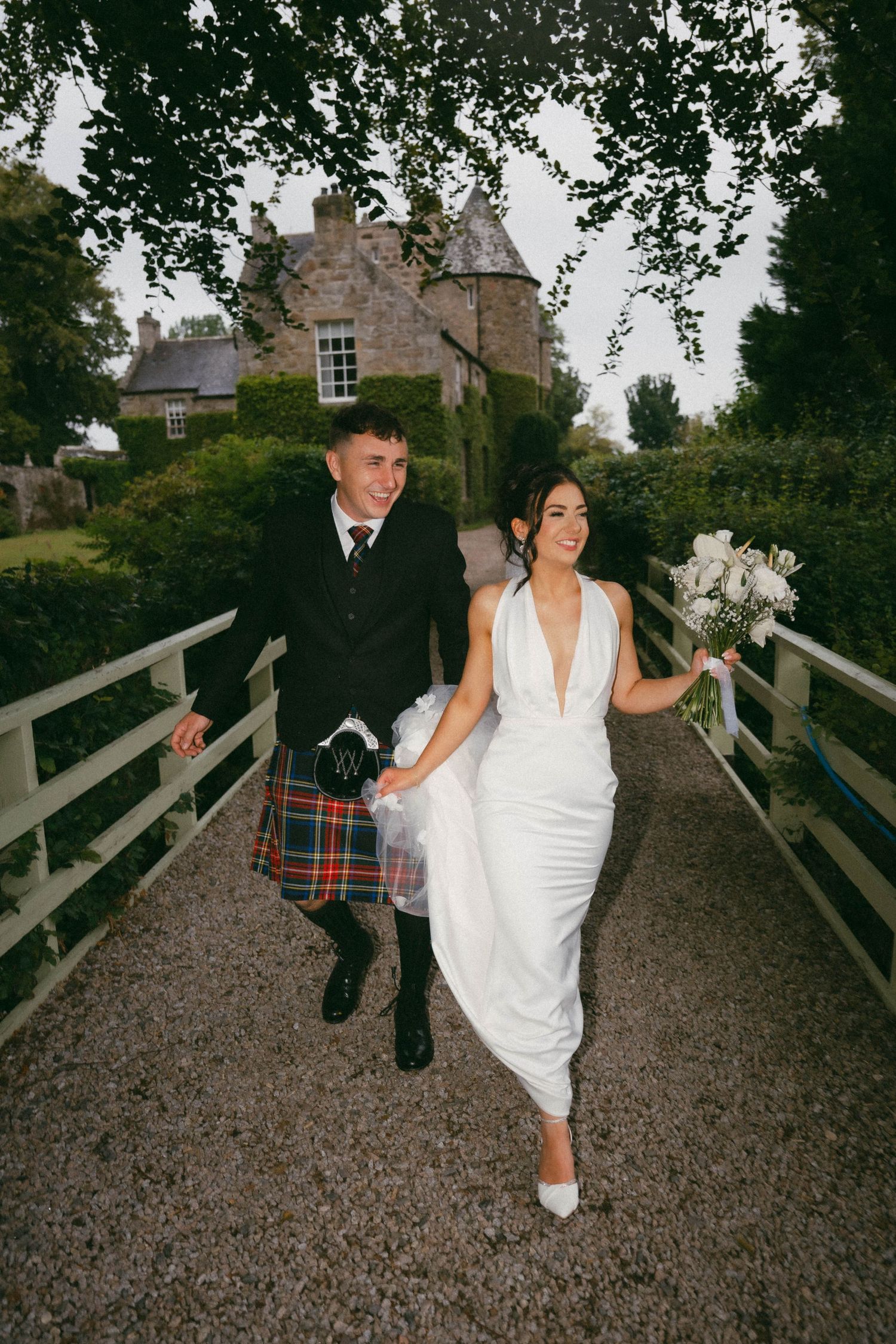 A couple walks across a bridge path with a Scottish castle in the background, the groom in a kilt and bride in white dress.