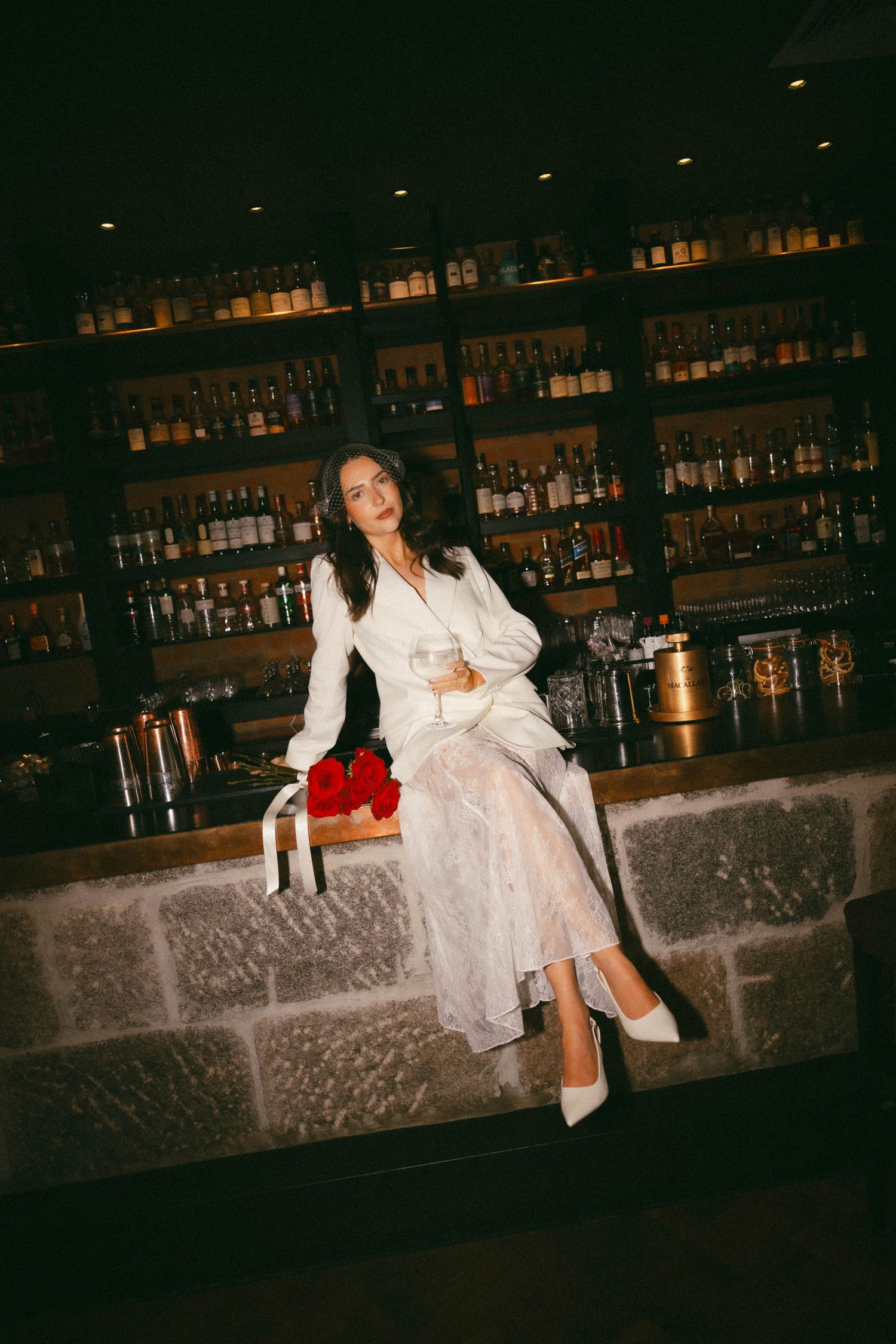 A woman in a white dress sits on a stone ledge at a bar with well-stocked shelves of liquor bottles in the background.