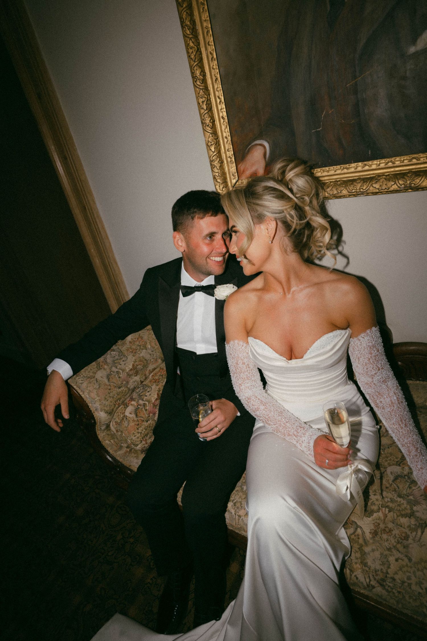 A couple shares an intimate moment on an ornate couch, the bride in a white gown and groom in classic tuxedo.