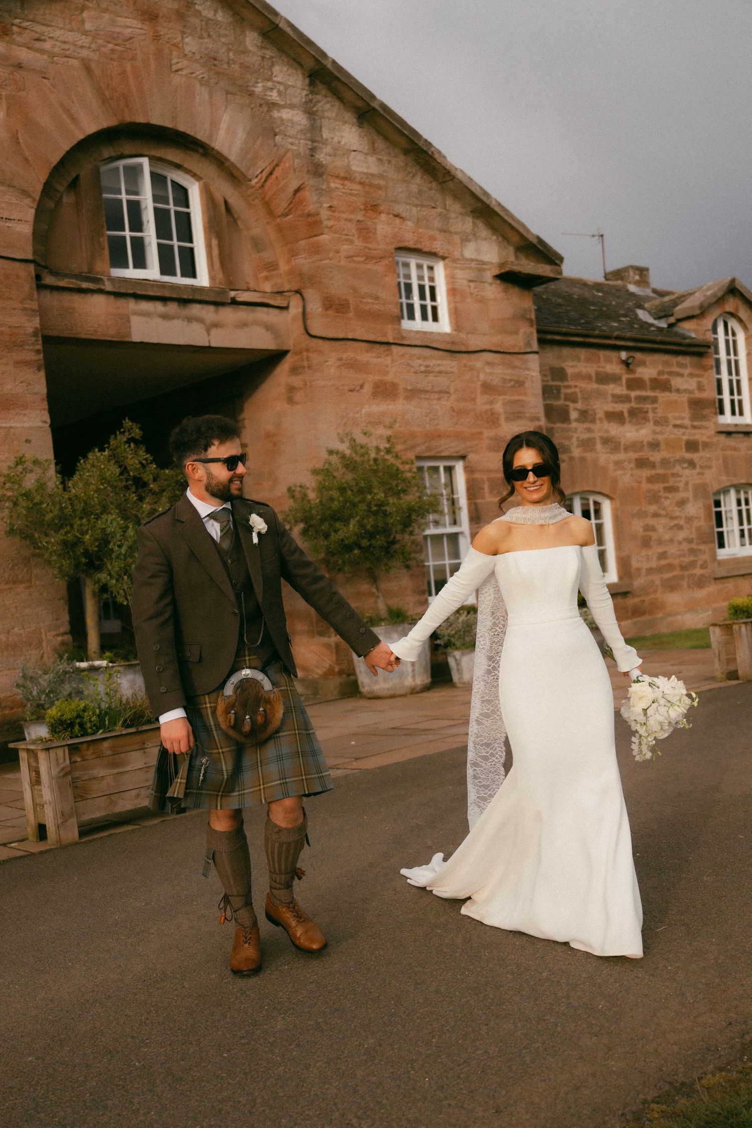 A bride and groom in Scottish wedding attire walk hand-in-hand outside a rustic stone venue.