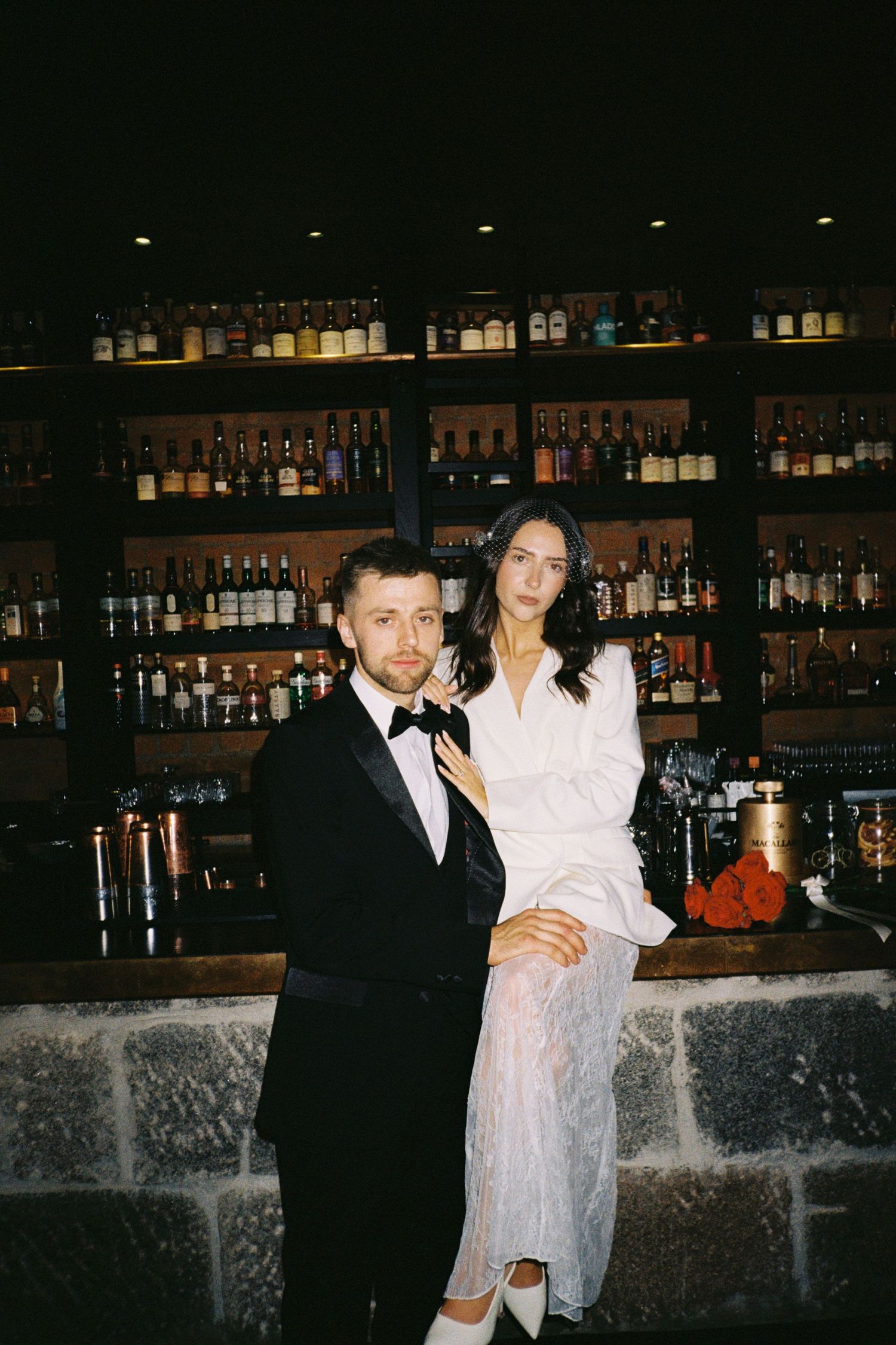 A couple poses together at a dimly lit bar with bottles displayed on shelves in the background.