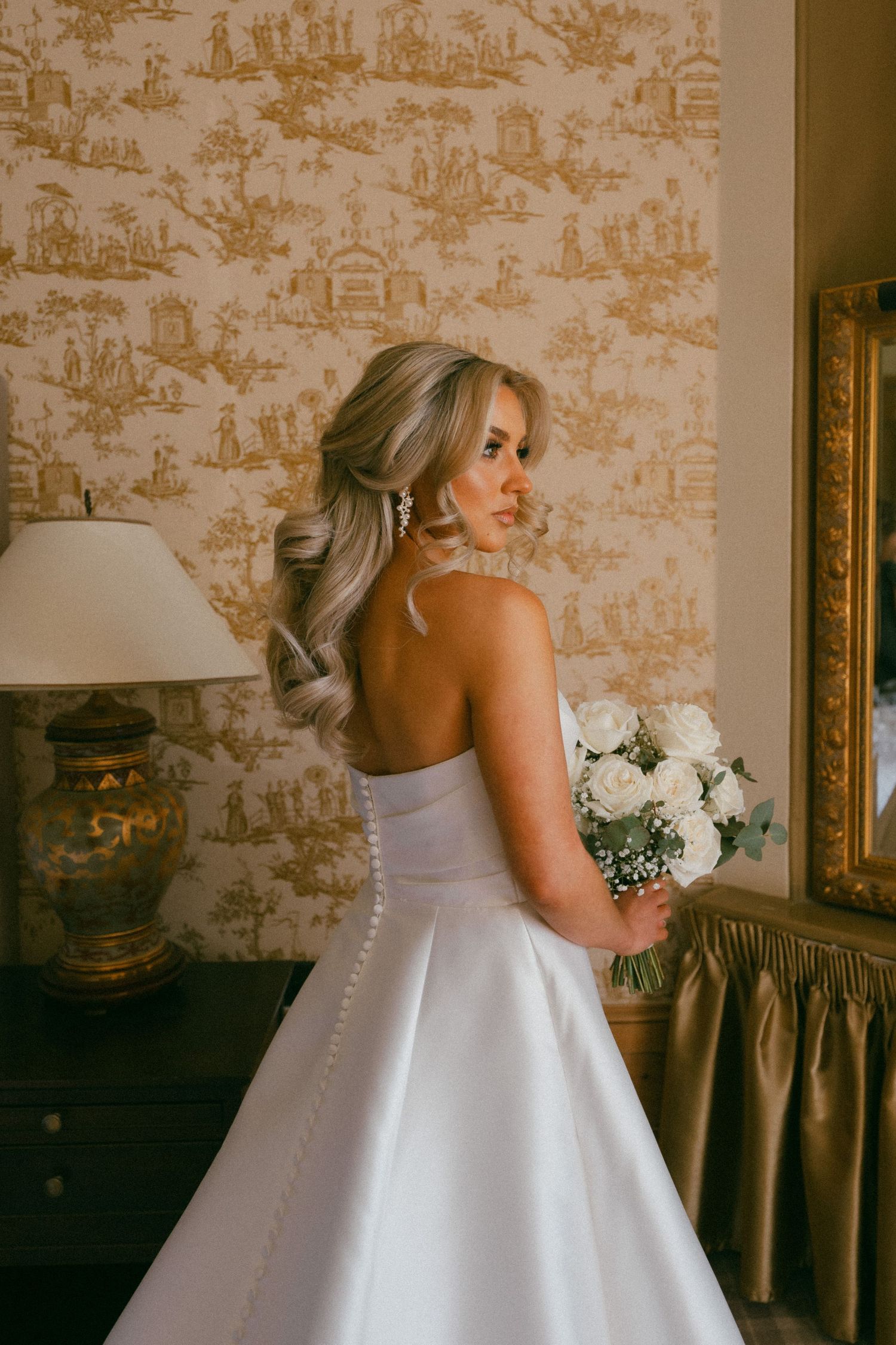 A bride in a classic white ball gown stands in an elegant room with gold damask wallpaper and vintage decor.