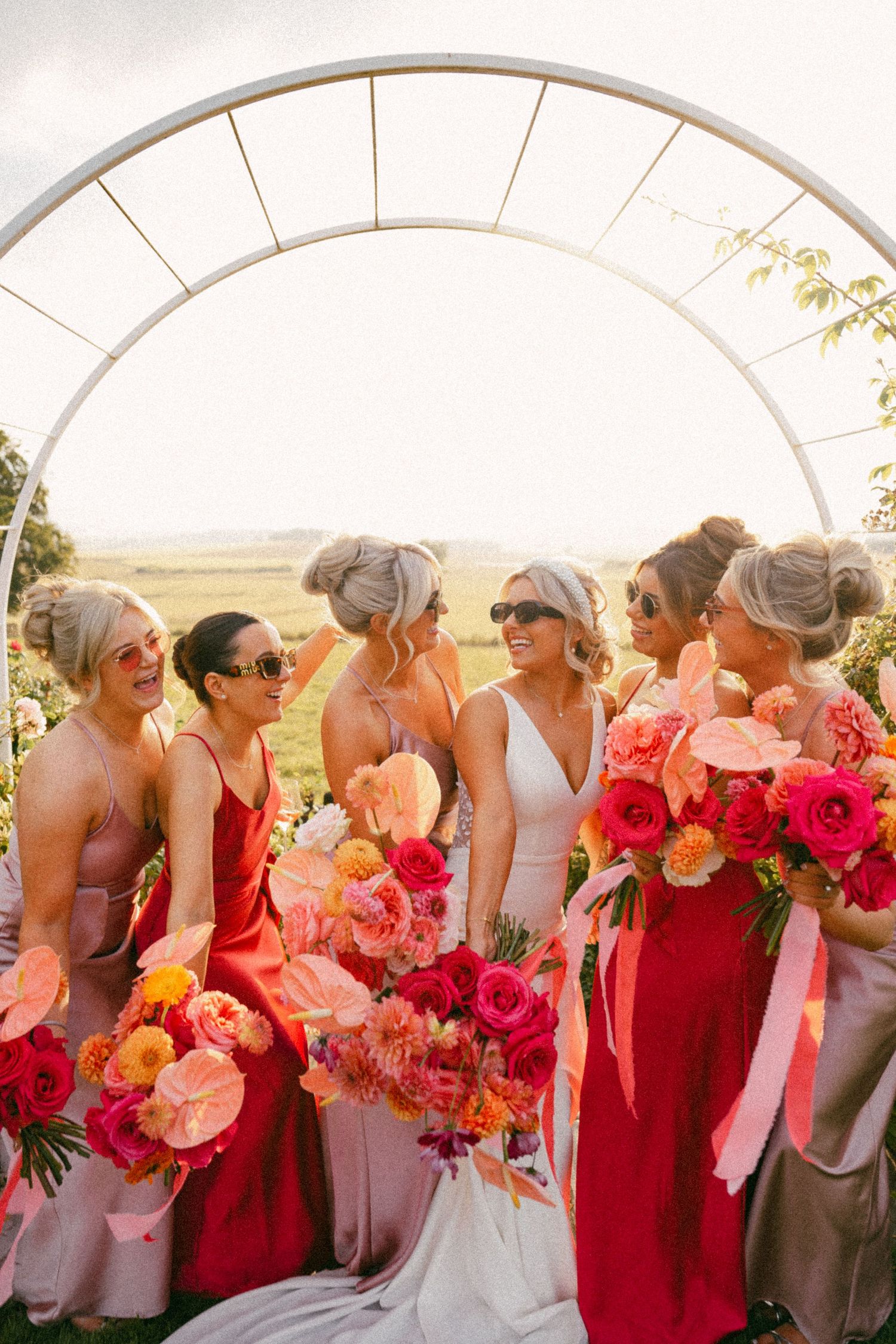 Bridesmaids in coral and pink dresses gather under white arch holding vibrant bouquets. iconic bridesmaid portraits, Barra barn
