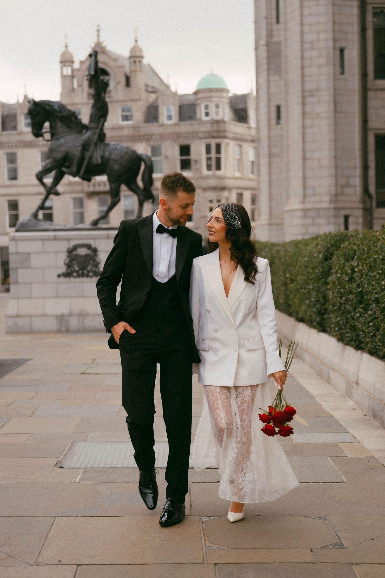 A couple in wedding attire strolls together near a stately building with ornate stone architecture.
