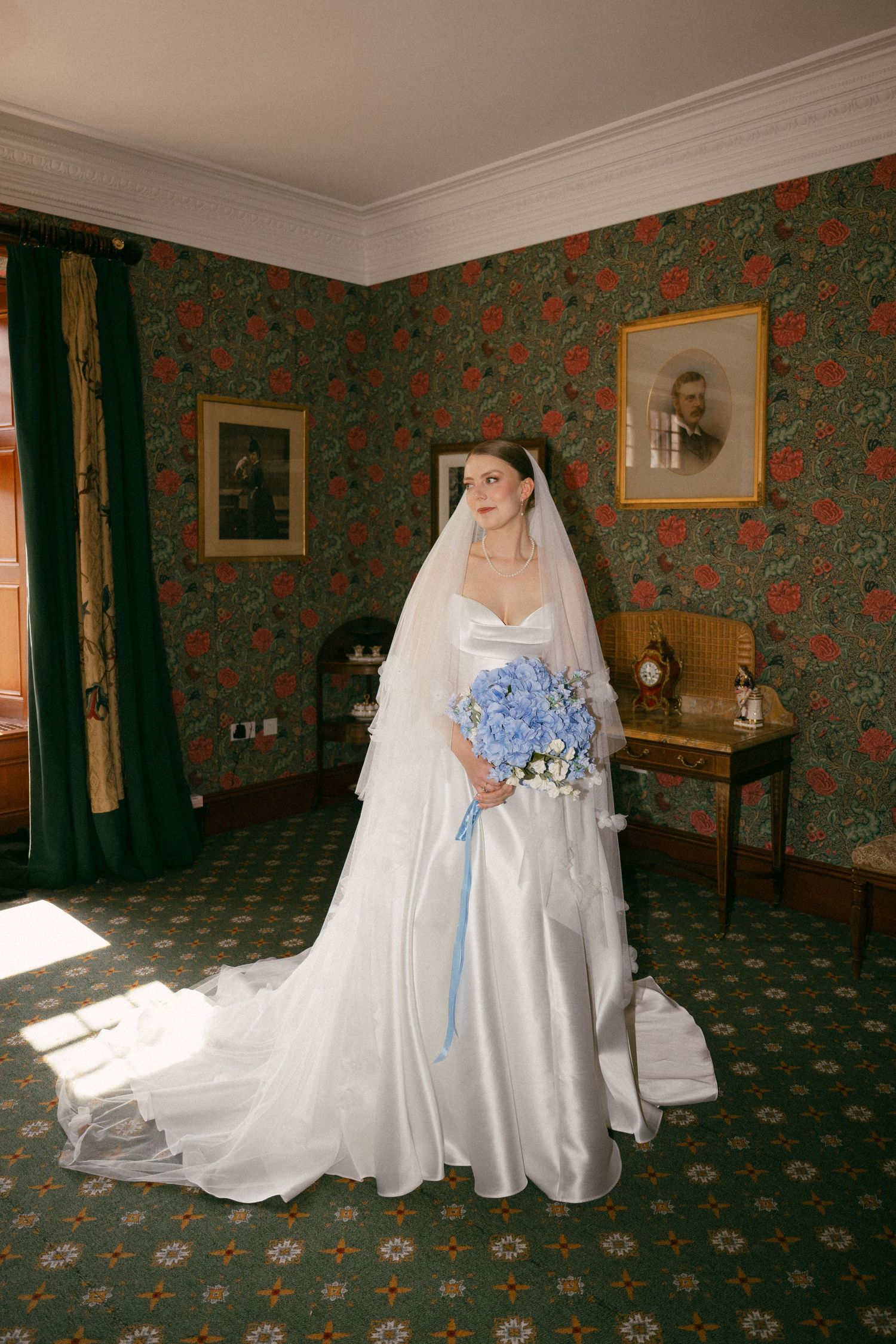 A bride poses in a classic white wedding gown with blue bouquet against floral wallpaper. Mar Lodge Estate Wedding
