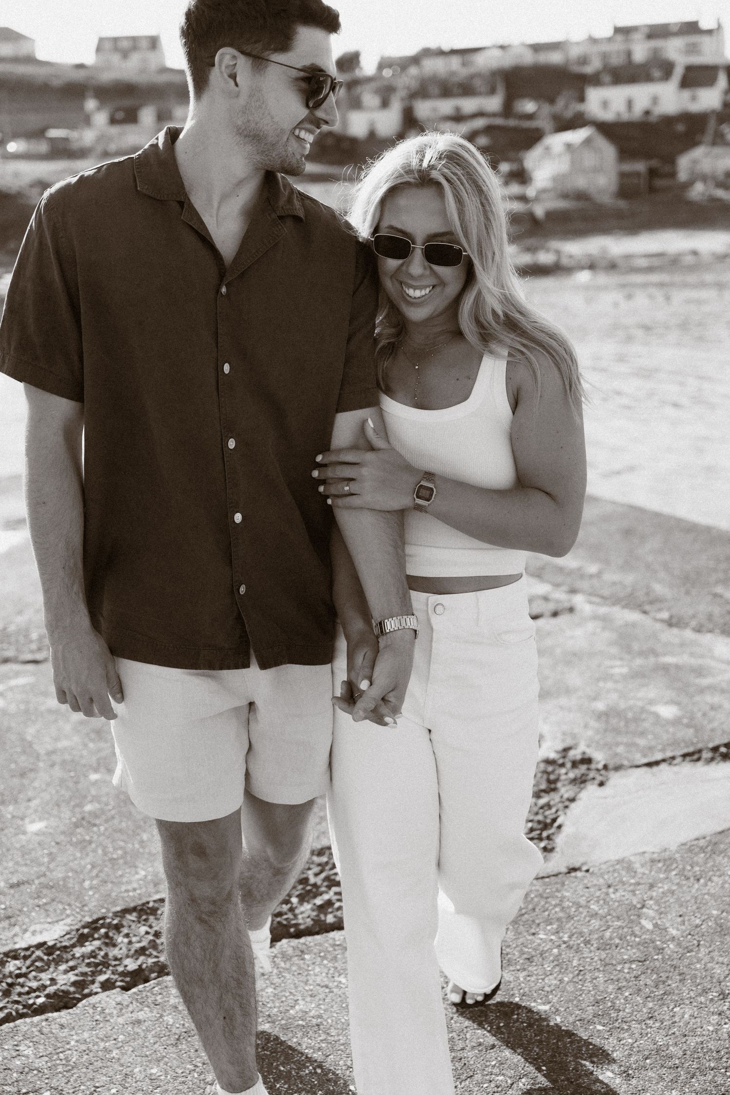 Black and white photo of a smiling couple holding hands and walking near the ocean on a sunny day.
