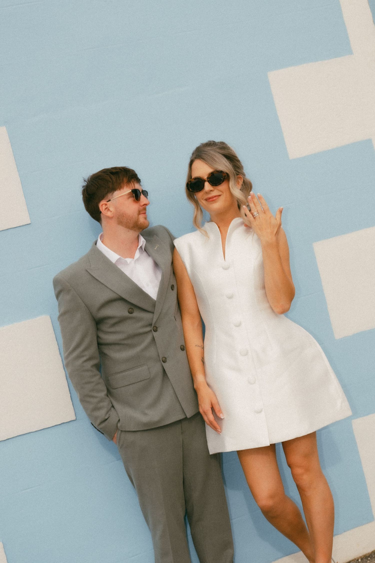 Couple in sunglasses posing against blue checkered wall, man in gray suit, woman in white mini dress showing ring.