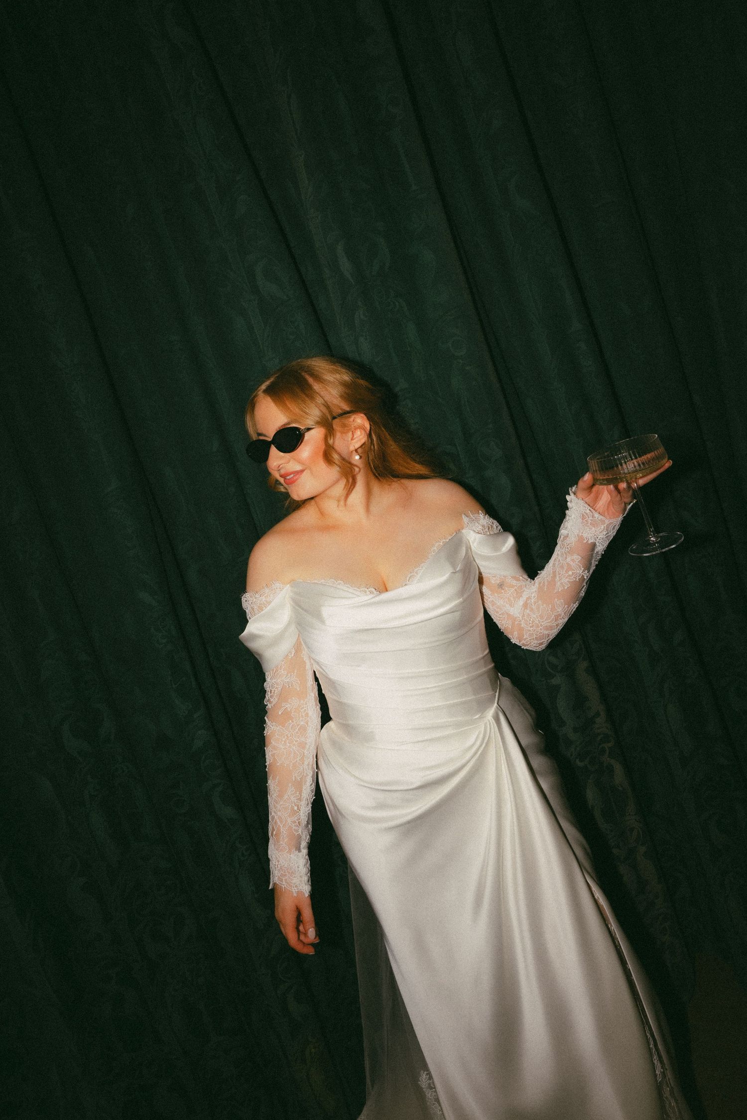 A bride in a white off-shoulder wedding gown with lace sleeves poses against a dark backdrop.