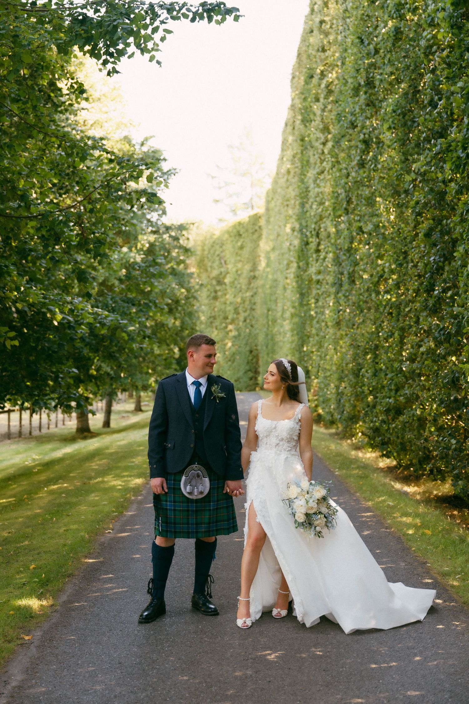 A couple poses on a tree-lined path during their Scottish wedding, with the groom in a kilt and bride in white dress.