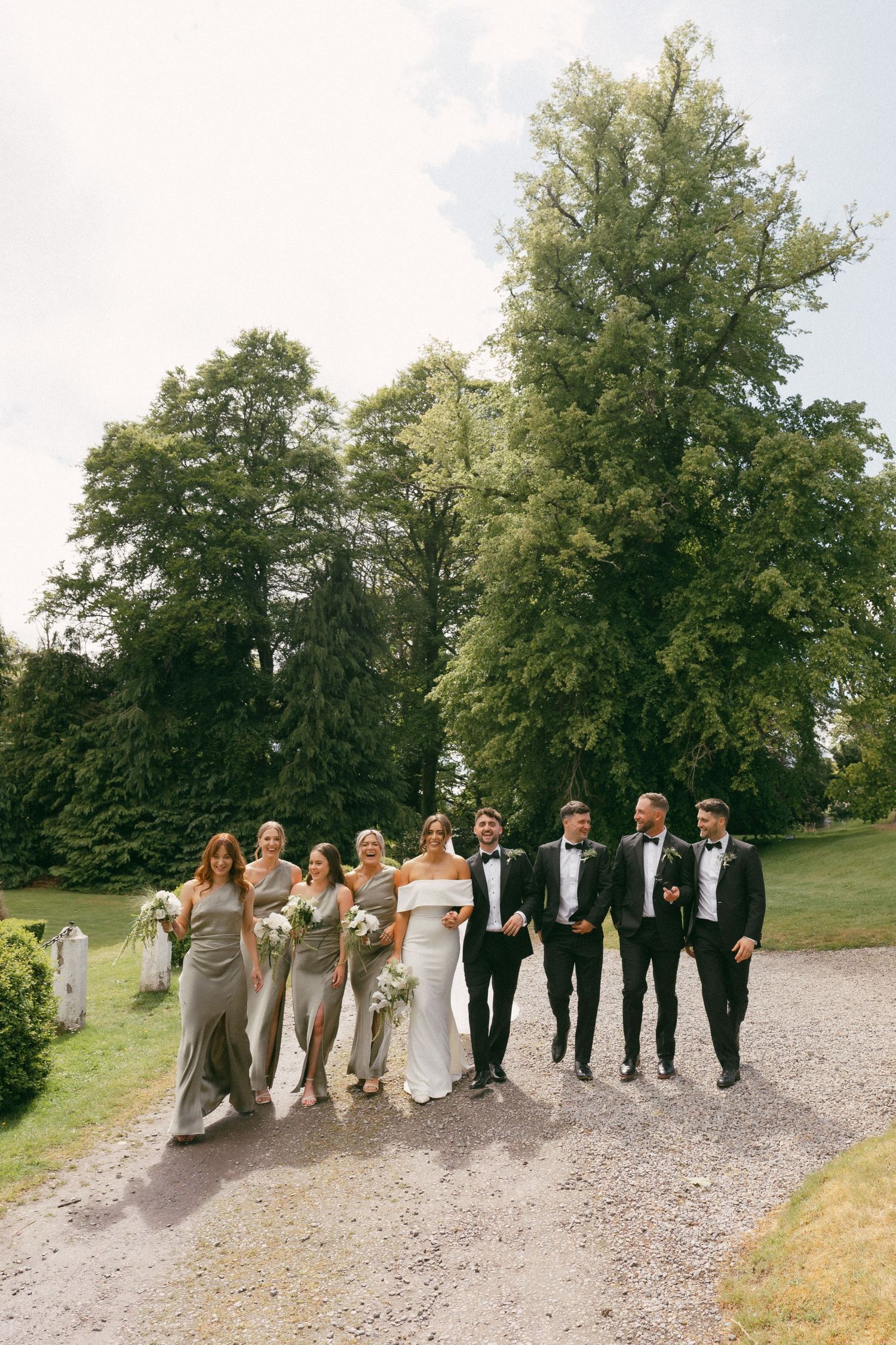 Wedding party in sage green bridesmaids dresses and black tuxedos walking together on a gravel path surrounded by trees.