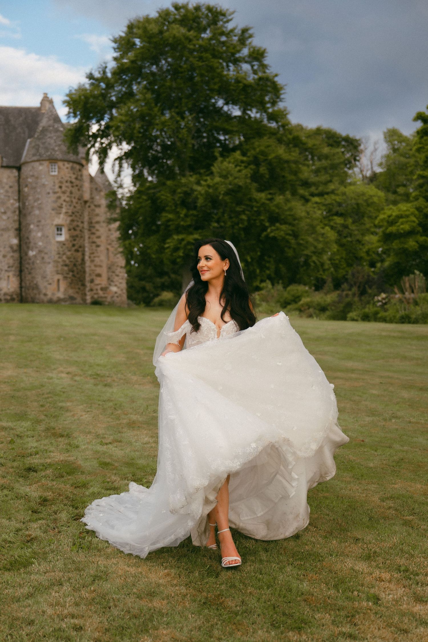 A flowing white wedding dress billows in the breeze on a grassy hill with an old stone castle in the background.