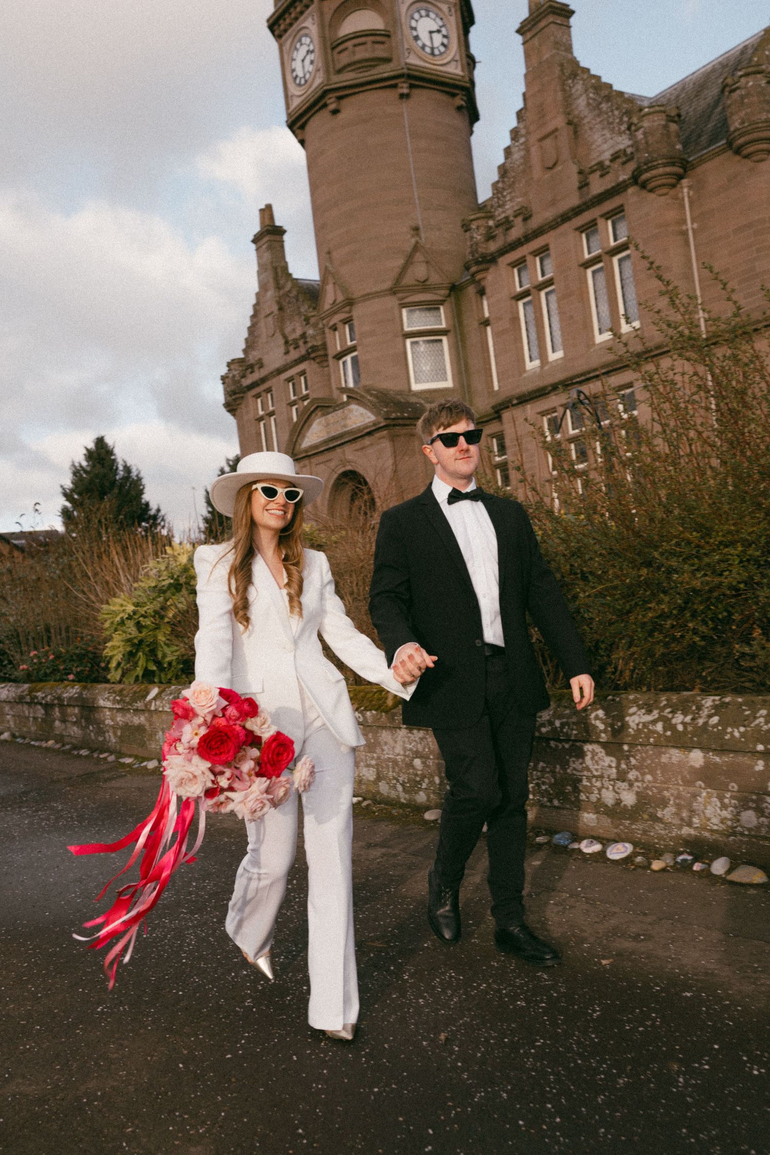 A couple in chic wedding attire walks along a path by a clock tower building. Aberdeen wedding photographer, wedding car elopement
